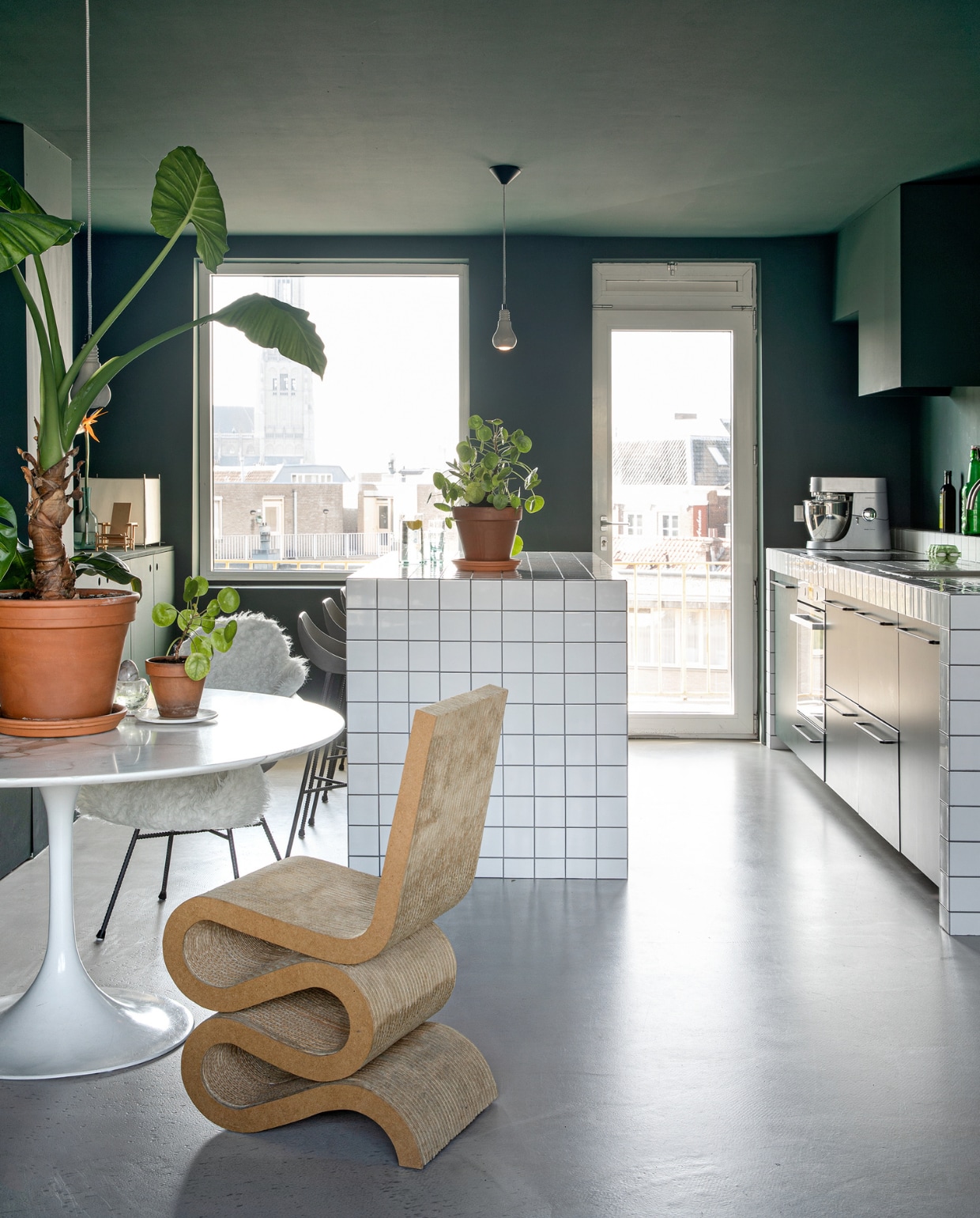 Keukeneiland van waterafstotend mdf met tegels van Hornbach. Vintage tafel Tulip van Eero Saarinen voor Knoll Studio. Planten en potten Intratuin. Betonnen hanglampen Edison van Tove Adman via Anno Design. Wiggle Side Chair van Frank Gehry is een erfstuk. Betonnen stoel en barkrukken Hauteville van Lyon Béton via La Boutique Blanche. Schapenvacht Ikea. Lamp Lotek van Benno Premsela is een erfstuk. Keukenmachine Kenwood. Verfkleur RAL 6012 zwartgroen.