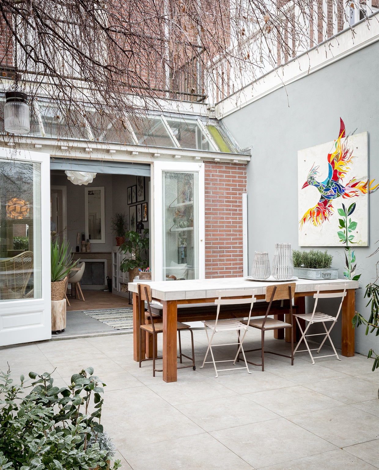 Muur buiten geverfd met buitenverf van de bouwmarkt in kleur gelijk aan vtwonen Ash. De eettafel (privébezit) is betegeld met tegels Neo Dessin Ambre uit de vtwonen-collectie by Douglas & Jones, 20×20 cm per m² € 69,50, bestrating tegels Hormigon Grey uit de vtwonen-collectie by Douglas & Jones, 60×60 cm per m² € 49,60 (www.vtwonen.nl). Op de buitentafel: stalen bak € 20,- (www.neeflouis.nl). Rozemarijn per m² (www.greenlifestylestore.nl). Antiek dienblad aluminium small € 115,-(www.householdhardware.nl). Lantaarn Bars van House Doctor, 30 cm € 21,99, lantaarn Bars van House Doctor, 40 cm € 27,50 (www.vtwonen.nl). Bistrostoelen in de kleur Linen € 64,- (www.extravert.nl). Oude houten vintage stoel (privébezit), soortgelijke stoelen € 69,- (www.ibizaoutdoor.nl). Kussen op de bank is op maat gemaakt door Meta Poulain. Lichtsnoer van House Doctor, 10-delig zwart € 94,95 (www.vtwonen.nl). Overig privébezit. vtwonen tv verbouwen of ver
