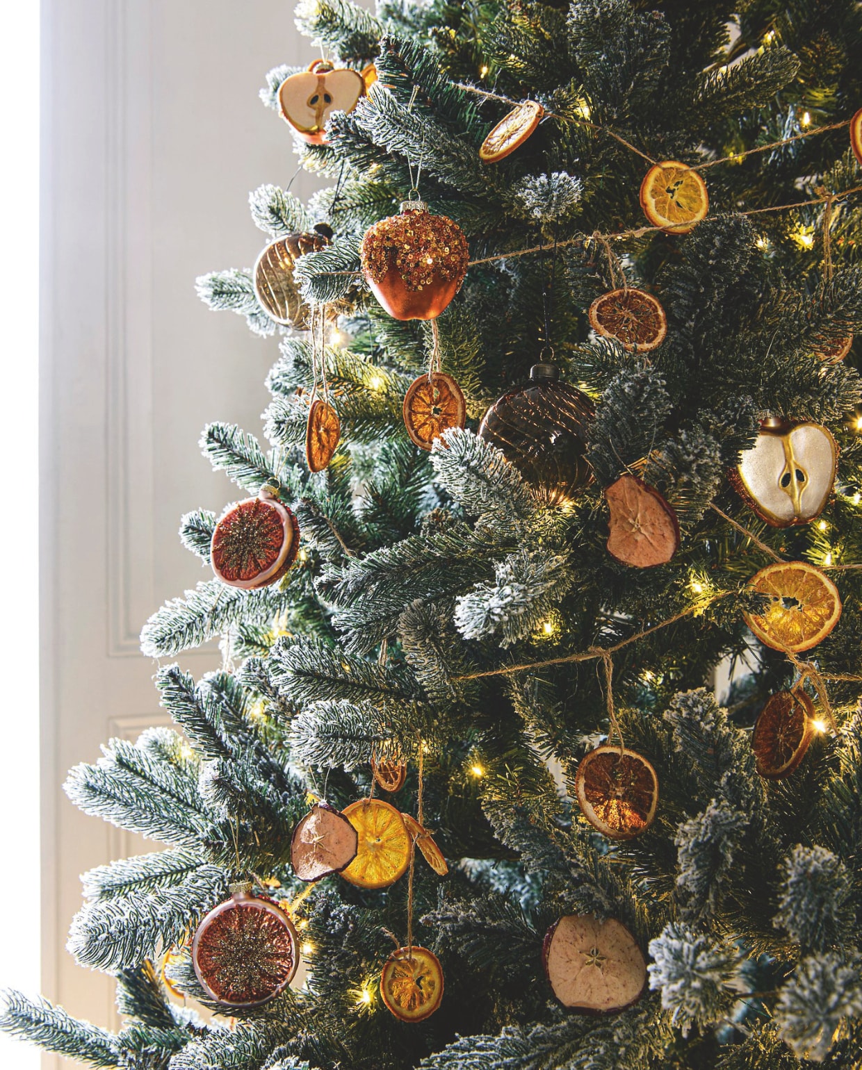 Kerstboom Anton Snowy, voor binnen en buiten, met ledlicht, 152x240 cm (Øxh) € 490,- (sirius.dk). Amberbruine glazen kerstbal van House Doctor € 5,49 (connox.nl). Glazen halve appel van House of Seasons € 4,-, glazen sinaasappel € 4,- (vtwonen.nl/shop). vtwonen 12-2024 | styling Marit Saladini | fotografie Louis Lemaire