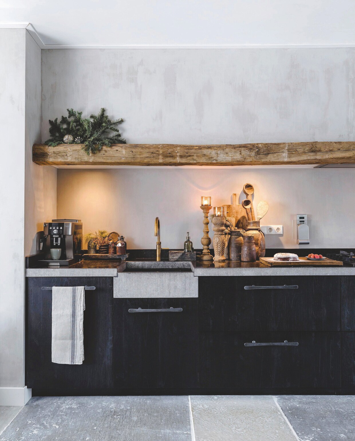 De keuken, van Keukenheeren Zwolle, is om de eikenhouten balk van Historisch Hout Haarle heen ontworpen. Het aanrechtblad en de gootsteen zijn van Belgisch hardsteen. De muur achter de keuken is behandeld met kalkverf en een coating. Alle accessoires op het aanrecht zijn van Rust &amp; tiek. vtwonen landelijk 07-2024 | Styling Rust &amp; tiek | Fotografie Corine Hoge Bavel-Jansen