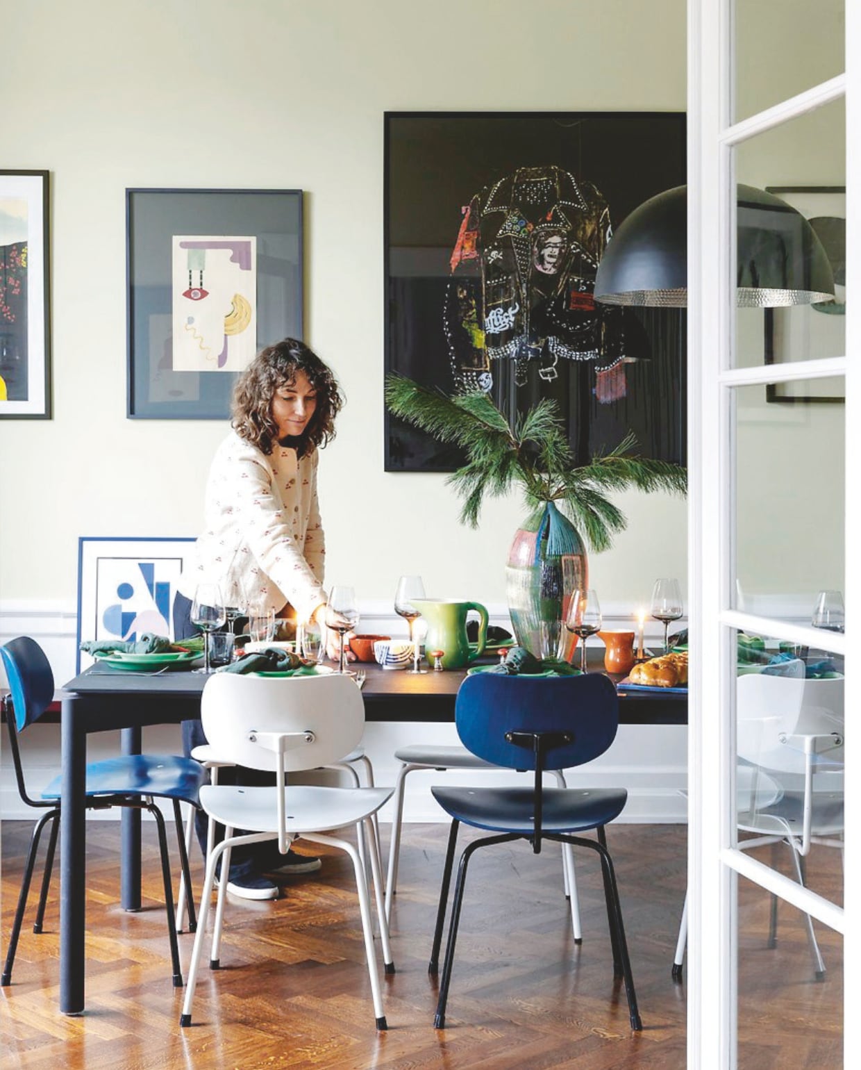 De eettafel van Out Objekte Unserer Tage uit Berlijn is gedekt voor kerstavond. Eetkamerstoelen Please Wait To Be Seated, hanglamp Mater, wandkunst van Jette Tosti Ibfelt en Rose Eken, borden en kan Ursula van Royal Copenhagen, Arne Jacobsen-bestek Georg Jensen, wijnglazen Brose, servetten Merci, kerstversiering vintage. vtwonen 12-2024 | styling Rikke Graff Juel | fotografie Christina Kayser