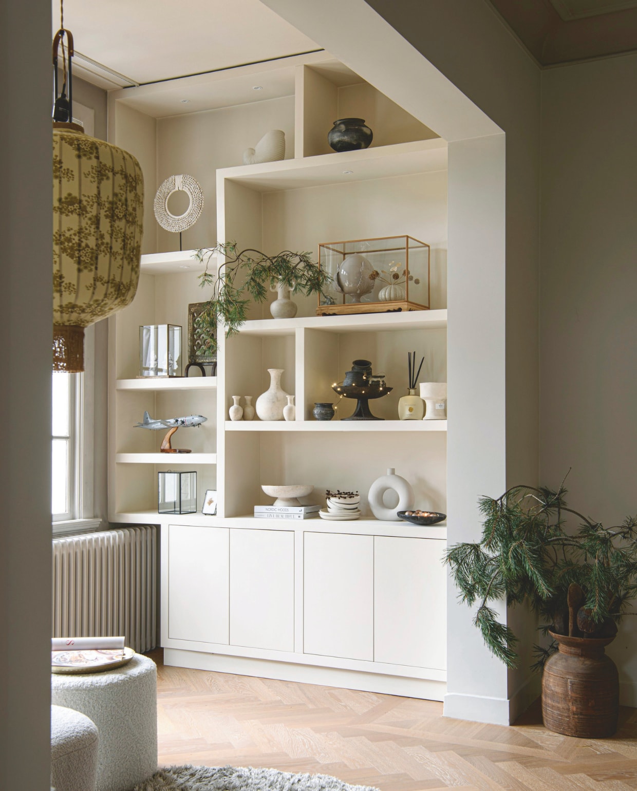 Vakkenkast op maat gemaakt. Poef Kwantum, vloerkleed Wehkamp, hanglamp HKliving. In vakkenkast: ornament Casa Krullenbol, vitrinekastjes Praxis, bloemist, Kwantum. Vazen, schalen, potten Household Hardware, Loods 5, Kwantum, lokaal gekocht en op reis. Vliegtuigmodel Orion cadeau gekregen, lijstje Sissy-Boy Homeland, geurstokjes Rituals. Kleur vakkenkast Dimity 2008 van Farrow &amp; Ball. vtwonen 12-2024 | styling Marit Saladini | fotografie Louis Lemaire
