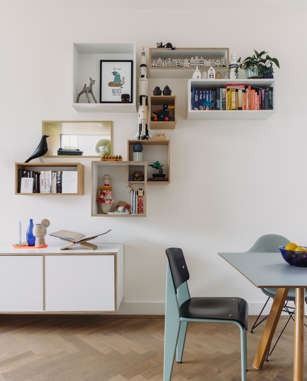 Stoel ontwerp Jean Prouvé voor Vitra, tafel en oranje dienblad Hay. Wandkast en kubussen gemaakt door meubelmaker. Boekenstandaard Loods 5, kandelaar en vaas The Clay Shaper, fles Kringloop. In vakken van boven naar beneden, v.l.n.r: hertje Marga van Oers, print kat Loods 5 Kunst, houten masker erfstuk, houten kat souvenir, huisjes KLM, huisjes op kastje zelfgemaakt en kringloop, fles Absolut Vodka, mintgroene bloempot erfstuk, raket en trekeend Lego, kopjes Cor Unum, typemachine Lego, discobal Marktplaats, vogel Vitra, houten bus Kringloop, porseleinen kunstwerk sample van Mickey Philips, bonsai Lego, gestreept vaasje Kringloop, Kokesho-pop souvenir. vtwonen DIY-special 11-2024 | fotografie Eva Bloem