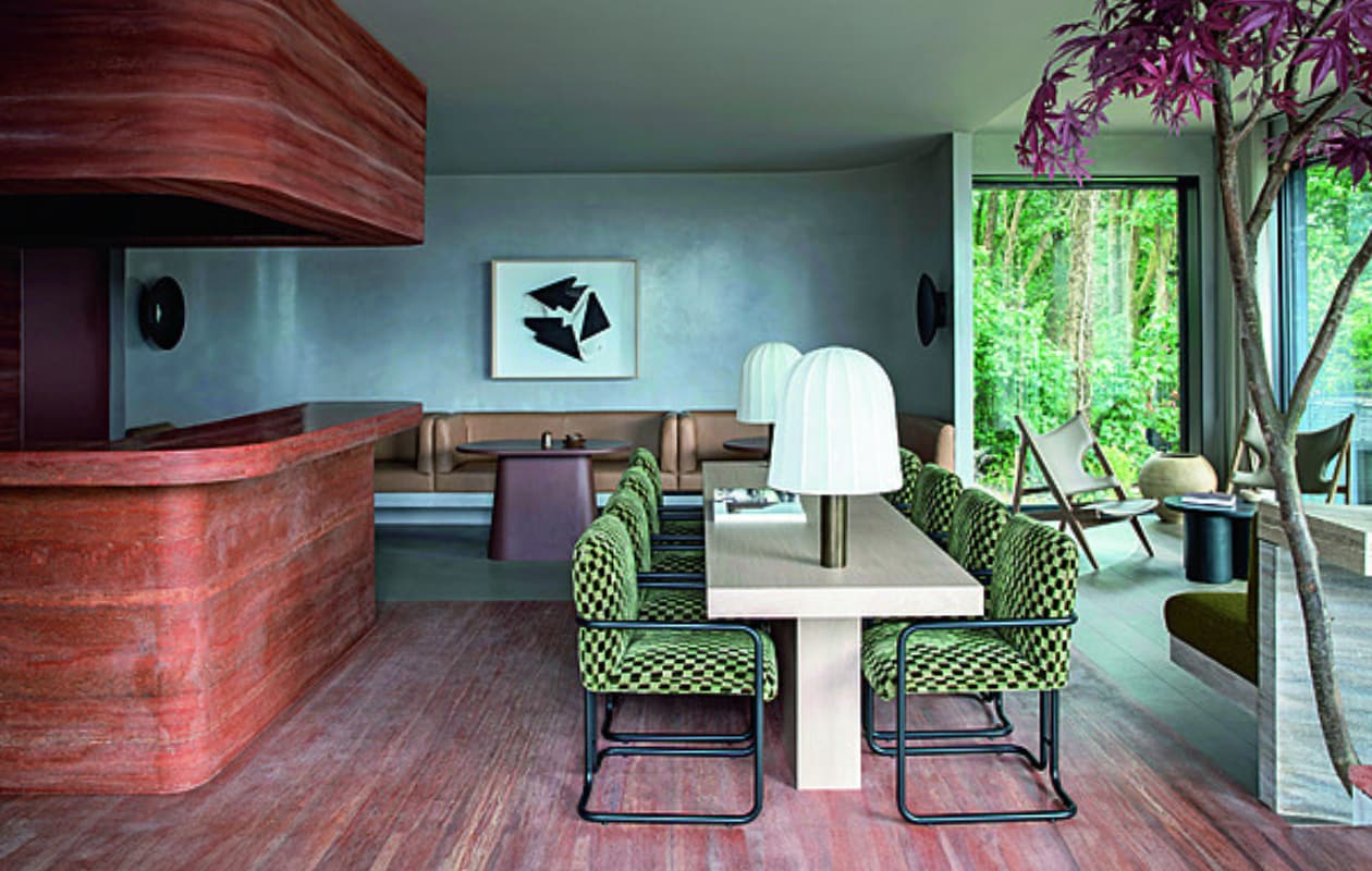 Het Clubhouse voor de gasten van de Woodland Suites met centraal een eiken tafel. Op de achtergrond op maat gemaakte leren banken en roodbruine tafels. Kunstwerk van Manolo Ballesteros. Voor het raam staat de Knitting Lounge Chair van Audo Copenhagen. Stijlvol Wonen 01-2025 | productie Living Inside | fotografie Ruth Maria Murphy