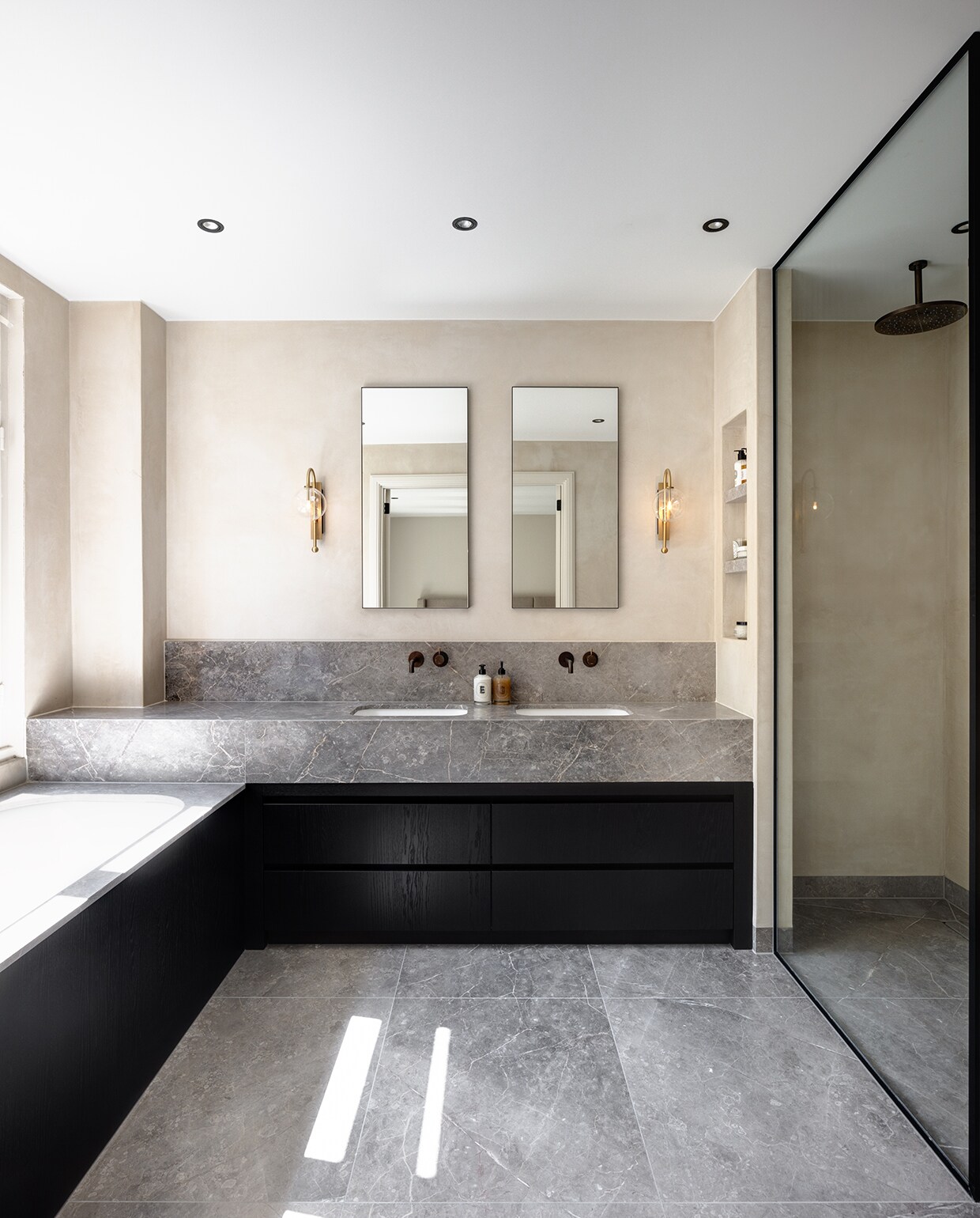 Vloer en bovenkant badombouw en badmeubel van Fior di Bosco-marmer, dat iets donkerder is dan het marmer beneden. Céline van Haaren Interiors ontwierp en realiseerde het eikenhouten badmeubel. Wandlampen Schwung. Bronzen kranen Hotbath. stijlvol wonen 2-2025 | Fotografie Jurrit van der Waal | Tekst Yvonne van Galen