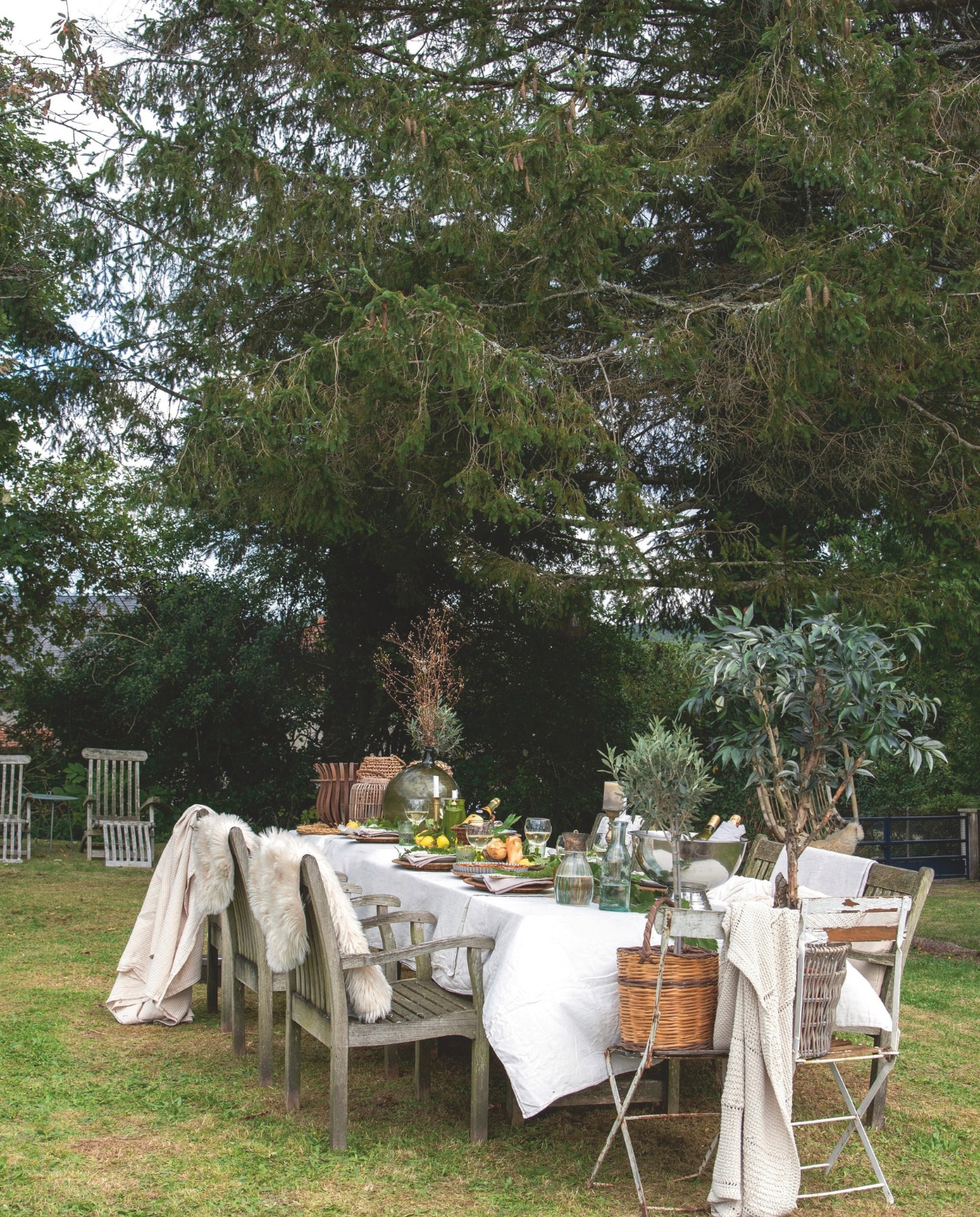 De grote teakhouten eettafel met stoelen in de schaduw van de spar biedt voldoende plaats aan de hele familie. De groene tafeldecoratie harmonieert perfect met de groentinten van de tuin. De deken haakte Loes zelf. vtwonen Landelijk 2-2025 | styling Peet Styling! | fotografie Joyce Vloet | tekst Marie Masureel