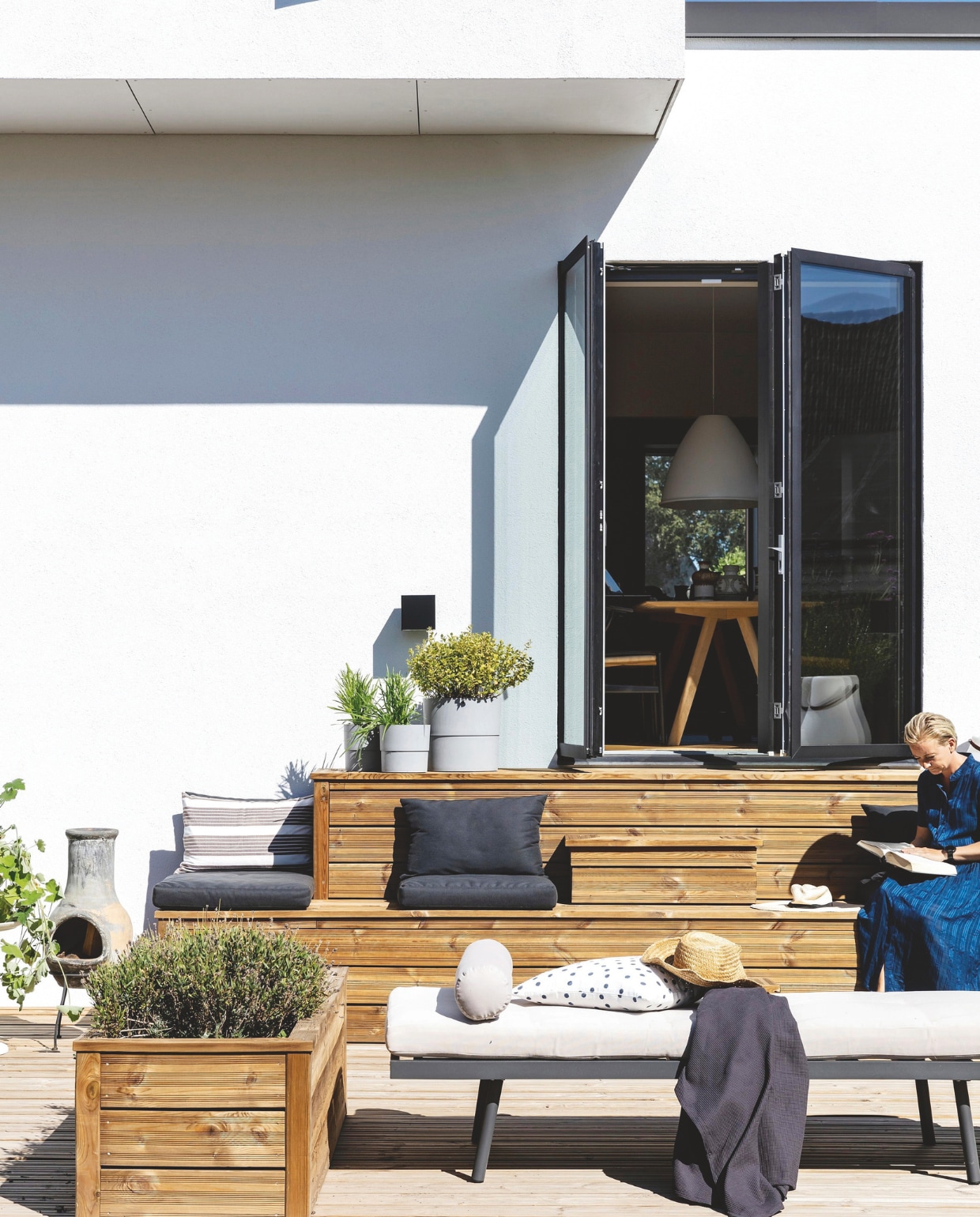 Het terras is gemaakt van geïmpregneerde houten planken. Hetzelfde hout is gebruikt voor de plantenbakken. Bank Bilka. Potten tuincentrum, met daarin onder andere buxusstruiken (Buxus sempervirens) en palmvarens (Cycas). Gestippeld kussen Ikea, andere kussens tuincentrum. vtwonen 01-2025 | productie living inside | styling en tekst rikke graff juel | fotografie christina kayser o. | tekst ina babbé