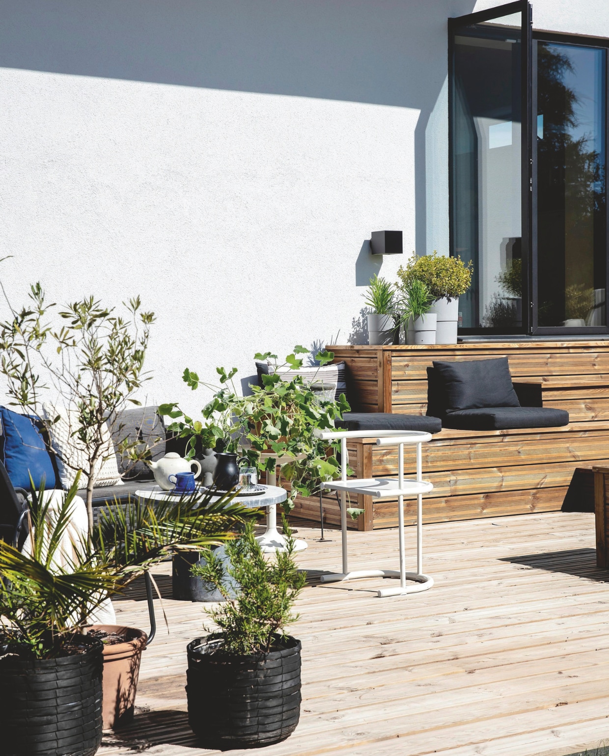 Naast de trap is de loungehoek, met een bank van Jysk en extra kussens van het tuincentrum. Betonnen tafel House Doctor. Stoel tweedehands. Plantenpotten kringloop en tuincentrum met daarin een olijfboompje (Olea europaea), buxus (Buxus sempervirens), lavendel (Lavandula angustifolia) en palmvaren (Cycas). vtwonen 01-2025 | productie living inside | styling en tekst rikke graff juel | fotografie christina kayser o. | tekst ina babbé