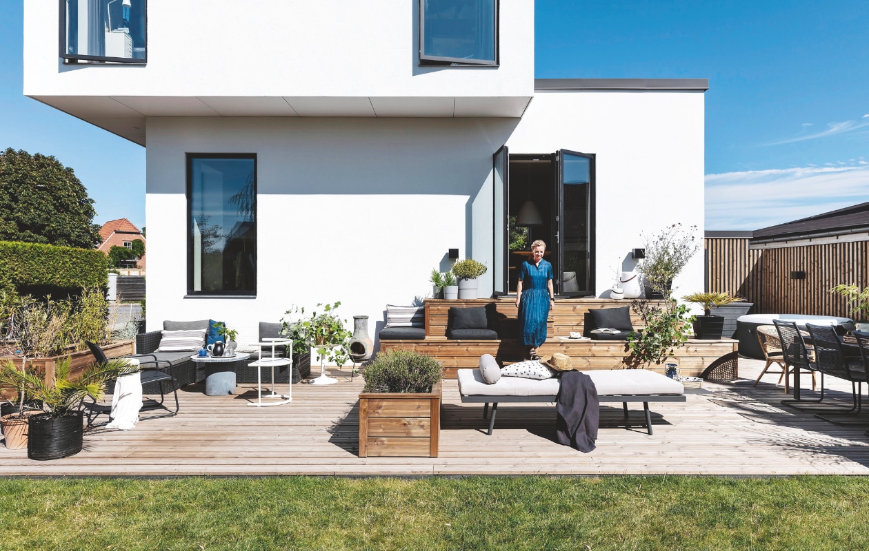 Hoekbank Jysk, kussens van het tuincentrum. Ronde betonnen tafel House Doctor. Witte stoel tweedehands. Plantenpotten kringloop en tuincentrum met daarin planten als olijfboompje (Olea europaea), buxus (Buxus sempervirens), lavendel (Lavandula angustifolia) en palmvaren (Cycas). In de potten aan weerszijden van de bank hanggeraniums (Pelargonium peltatum). Daybed Bilka. Stippelkussen Ikea. Buitenhaard van klei uit Mexico. Op de trap twee ledbuitenlampen van Nordicluxx. De grote heeft een ingebouwde speaker en kan ook gebruikt worden als wijnkoeler. Eettafel Sylvan, zwarte stoelen Ikea. vtwonen tuin 11 | productie living inside | styling en tekst rikke graff juel | fotografie christina kayser o. | tekst ina babbé