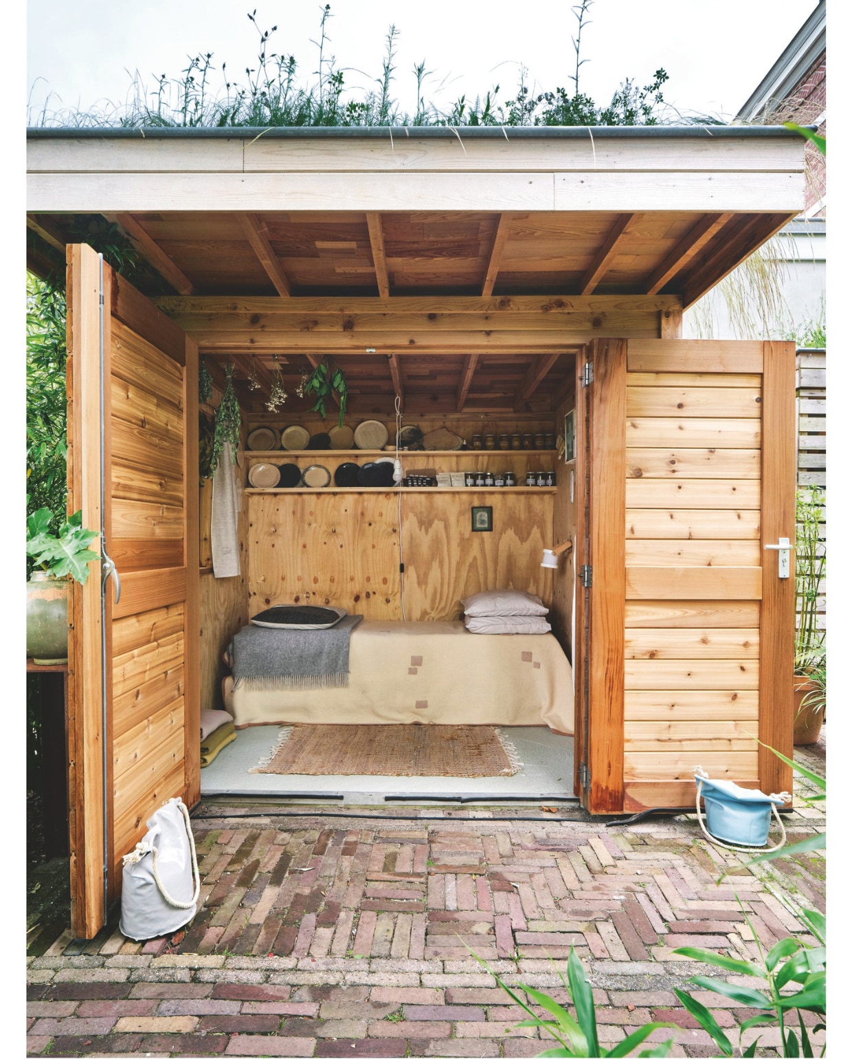 Tuinhuis eigen ontwerp. Bed van pallets, deken, gordijnen, kussens, lamp en servieswerk Brigit Vermeulen. Oplaadbaar harmonicalampje Go Living. Vloerkleed Zeeman. Kunstwerk en oud medicijnkastje kringloop. vtwonen 03-2025 | fotografie René van der Hulst