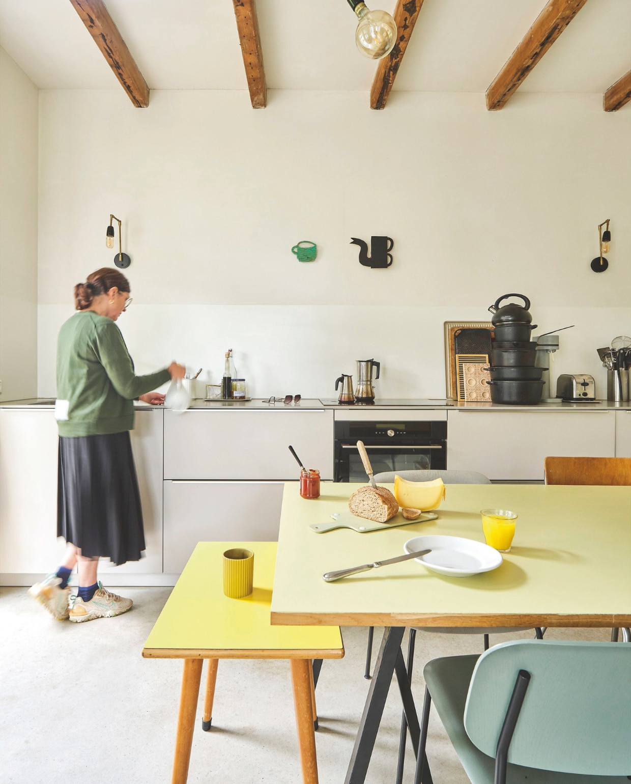 Keuken Bruynzeel. Eettafel gemaakt van schragen van Hay, tafelblad van de bouwmarkt, geverfd in Cornflower Field van Flexa Creations. Stoel Soft Edge van Hay, herkomst Belgische buisframestoelen onbekend. Kleine gele tafel kringloop. Werken aan de wand Klaas Gubbels. Wand- en plafondlampen Brigit Vermeulen. Achterwand keuken is gedeeltelijk zijdeglans gelakt met Ral 9010, rest is onbewerkt stucwerk. Pannen diverse merken, onder andere Le Creuset. Vloer gevlinderd beton met vloerverwarming.