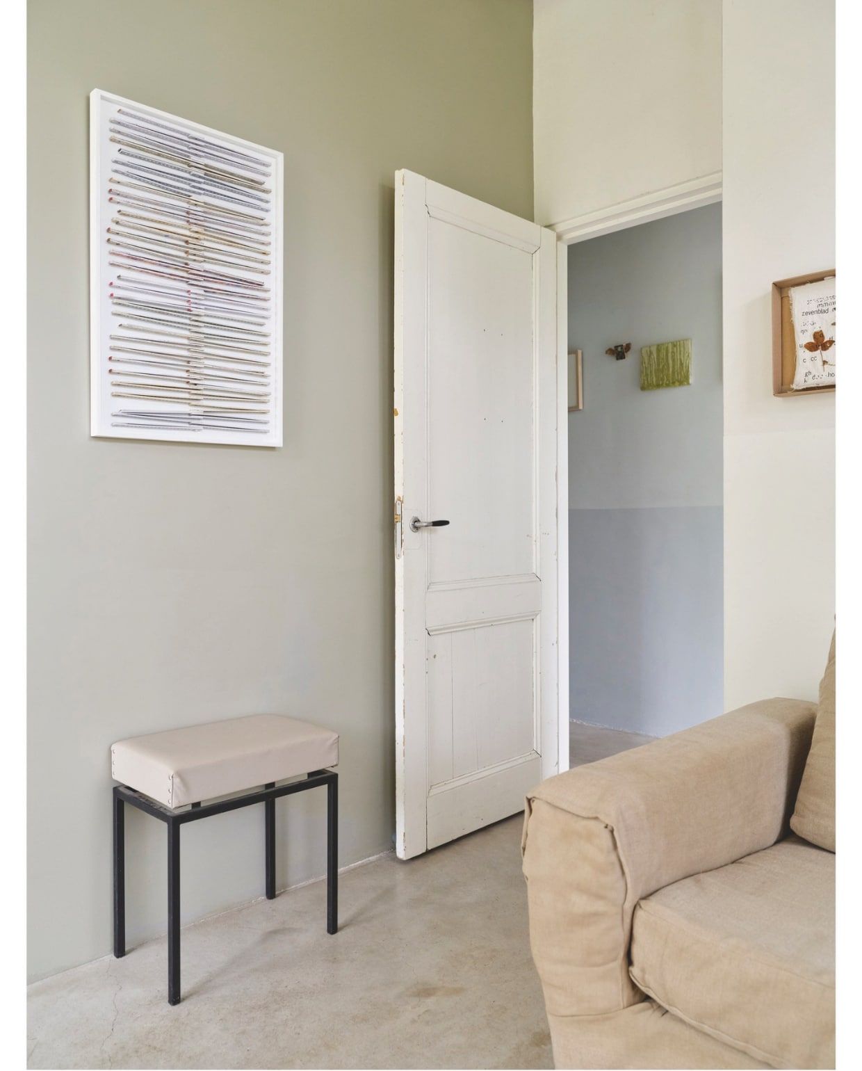 Bank kringloop, bekleed met linnen. Kruk kringloop, gespoten en bekleed met leer. Muurkleur woonkamer Portland Stone Dark, kleur gang Bone China Blue Pale van Little Greene. Kunstwerk woonkamer Ilse Vermeulen, werken  in de gang Ilse Vermeulen en Han Klinkhamer. Vloer gevlinderd beton. vtwonen 03-2025 | fotografie René van der Hulst