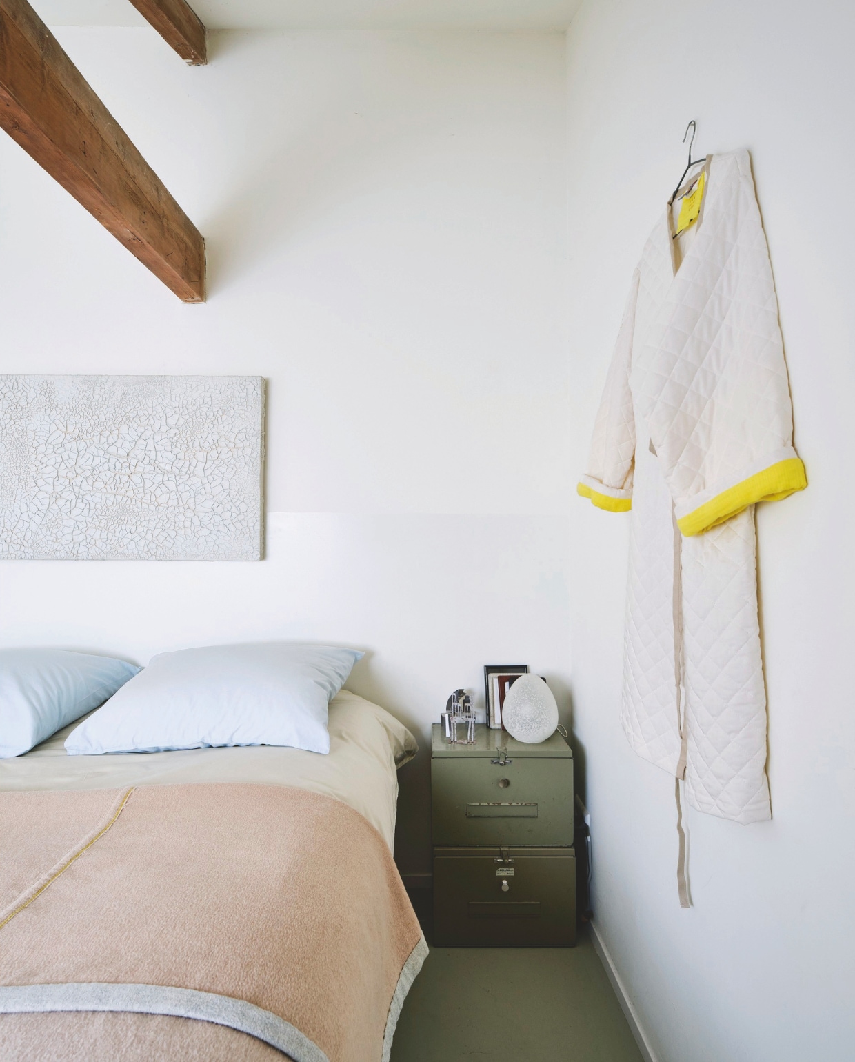 Beddengoed, deken, dekbedovertrek en kussenslopen Brigit Vermeulen. Jas Brigit Vermeulen voor Social Label. Schilderij gekregen. Legerkist uit vorige huis. Bedlamp Egg Shaped Glass Zo Goed als Oud. Wand achter bed gedeeltelijk gelakt in Ral 9010. Houten vloer geverfd in Ral 7032. vtwonen 03-2025 | fotografie René van der Hulst | tekst Merel van der Lande