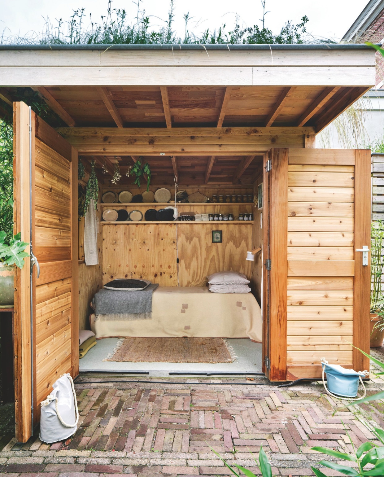 Tuinhuis eigen ontwerp. Bed van pallets, deken, gordijnen, kussens, lamp en servieswerk Brigit Vermeulen. Oplaadbaar harmonicalampje Go Living. Vloerkleed Zeeman. Kunstwerk en oud medicijnkastje kringloop. vtwonen 03-2025 | fotografie René van der Hulst | tekst Merel van der Lande