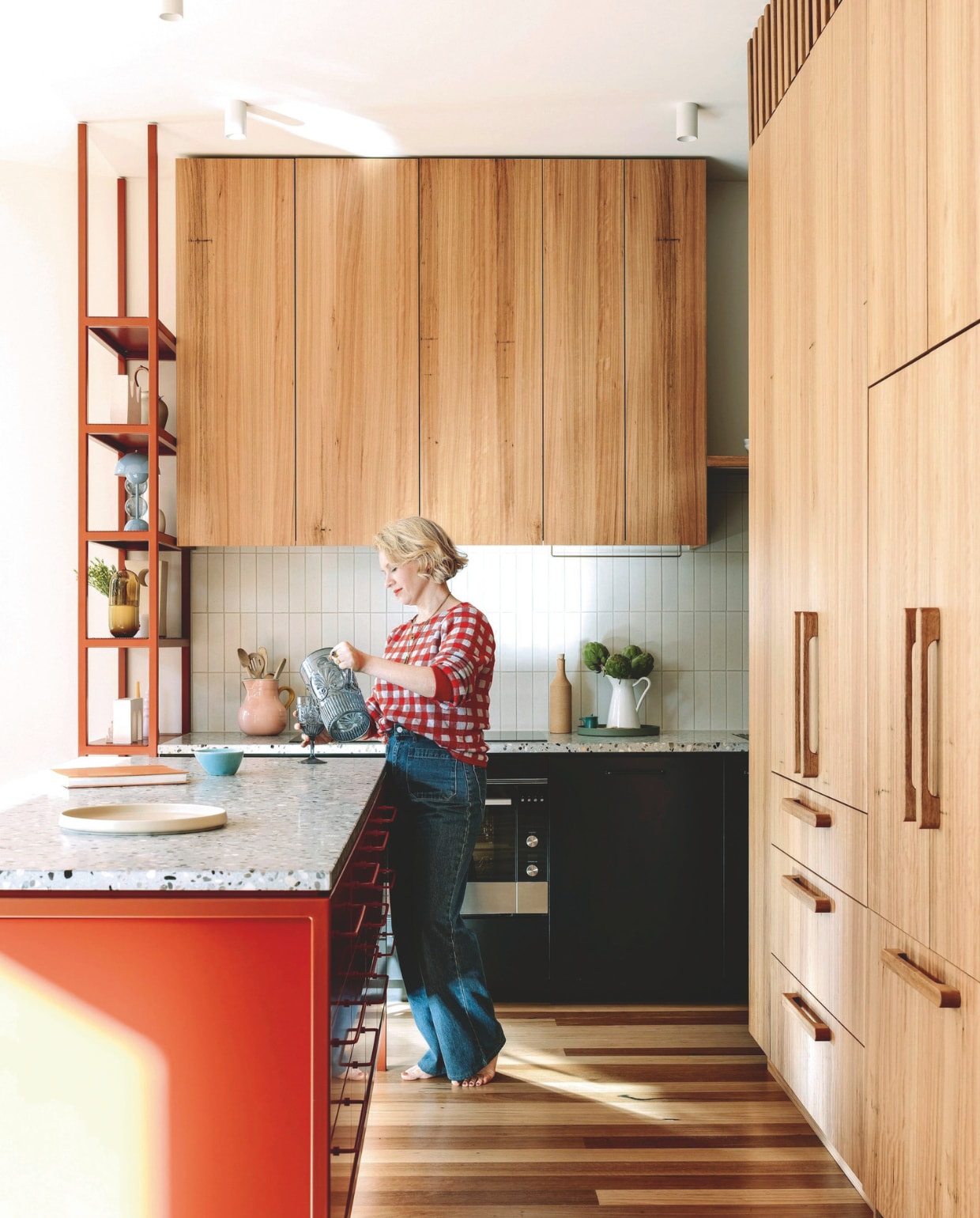 Keukenkasten, -eiland en hoge stellingkast, ontworpen door Imogen Pullar Architecture, gebouwd door J&amp;RC Builders met duurzame blackbutt-fineer. Handgemaakte houten handvaten Auburn Woodturning. Rood stalen frame en handgrepen Lounge van Kethy. Terrazzo keukenblad Moon Garden van Fibonacci. Tegels Hida van Academy Tiles. vtwonen 03-2025 | styling belle hemming | fotografie marnie Hawson | tekst caroline westdijk