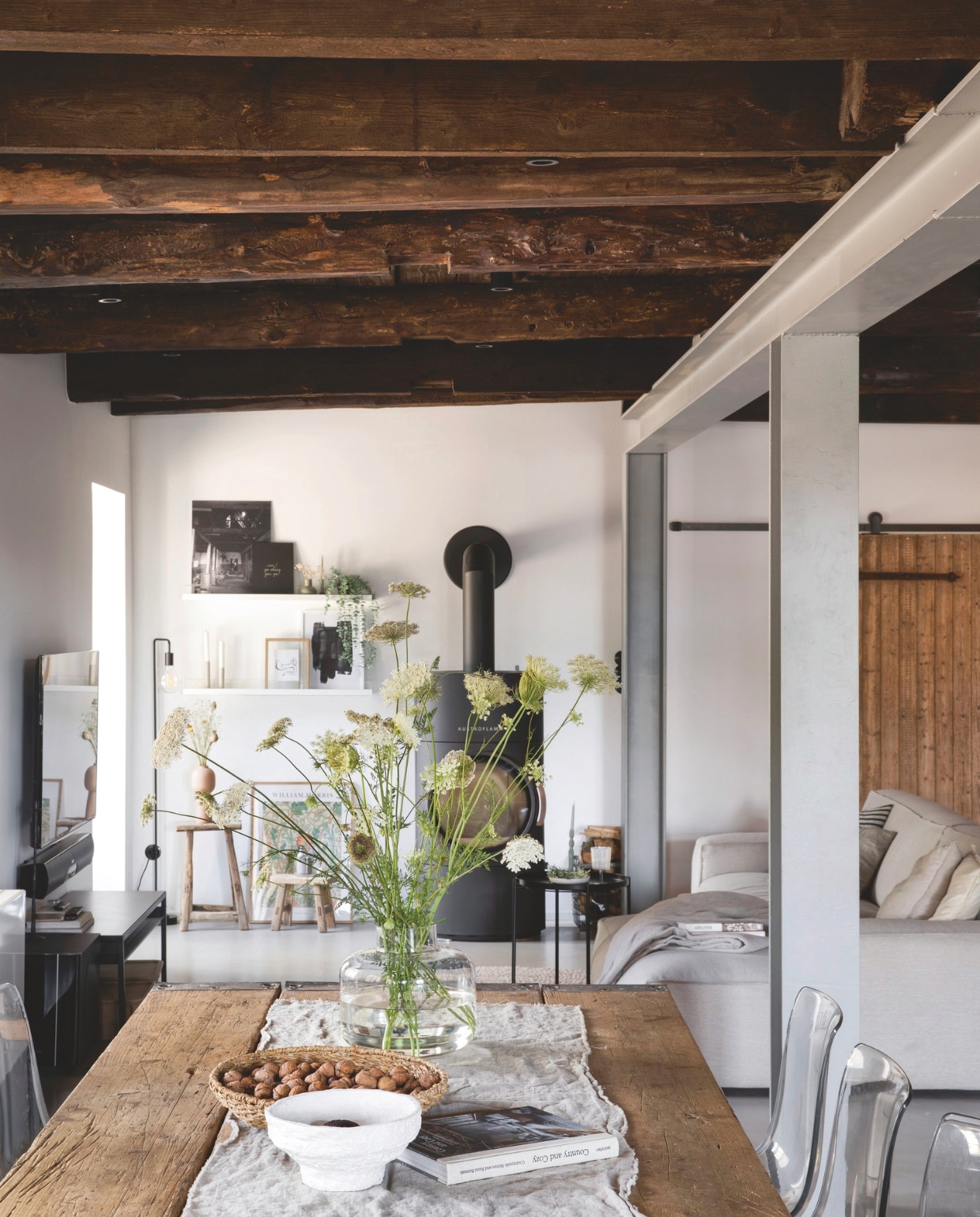 Eettafel van oude steigerplanken gemaakt door Jan. Kachel Austroflamm. Krukken Cooperz Wonen. Vloerlamp Leen Bakker. Lijsten Ikea. Bank Fést. Soortgelijke vaas Mica Decorations. De betonnen vloer lag er al, net als de stalen balken ter versteviging, die Anna-Lena grijs maakte. vtwonen Landelijk 03-2025 | Styling Miriam Hannemann | fotografie André Reuter | tekstbewerking Chris Muyres