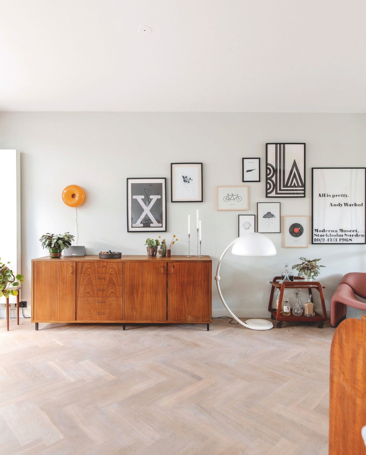 Dressoir vintage. Witte staande lamp Elio Martinelli uit ouderlijk huis Jessica. Barkast vintage. Oranje lamp Varmblixt van Ikea. Kandelaars Piet Hein. Bruine potten, notenschaal en groene plantenpot vintage. Kunst met de klok mee: grote X herkomst onbekend, ernaast Martin Moore, tandem gekocht op markt, daarboven gekocht op Schiermonnikoog, Bla poster One must Dash, schelp Inkylines, wolk Hyshil Sander, elpee gekocht op markt vtwonen 03-2025 | fotografie Studio Keeelly | tekst Caroline Westdijk