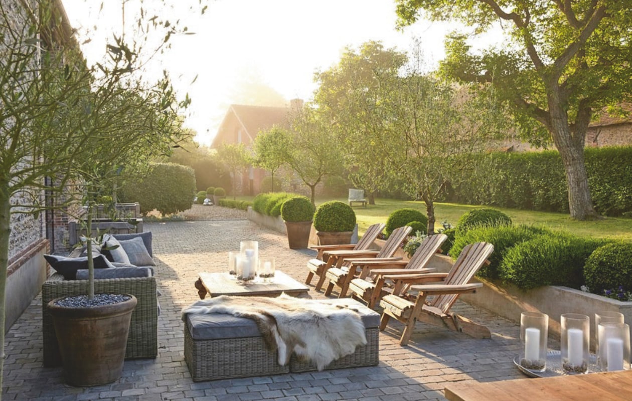 Buxusbollen, blauwe klokjes en lavendel  in de plantenbakken. Ligstoelen Kenas Home. Het terras is omlijst door stenen plantenbakken en olijfbomen in houten bakken. vtwonen landelijk 03-2025 | Styling José Spaans | Fotografie Sarah Van Hove