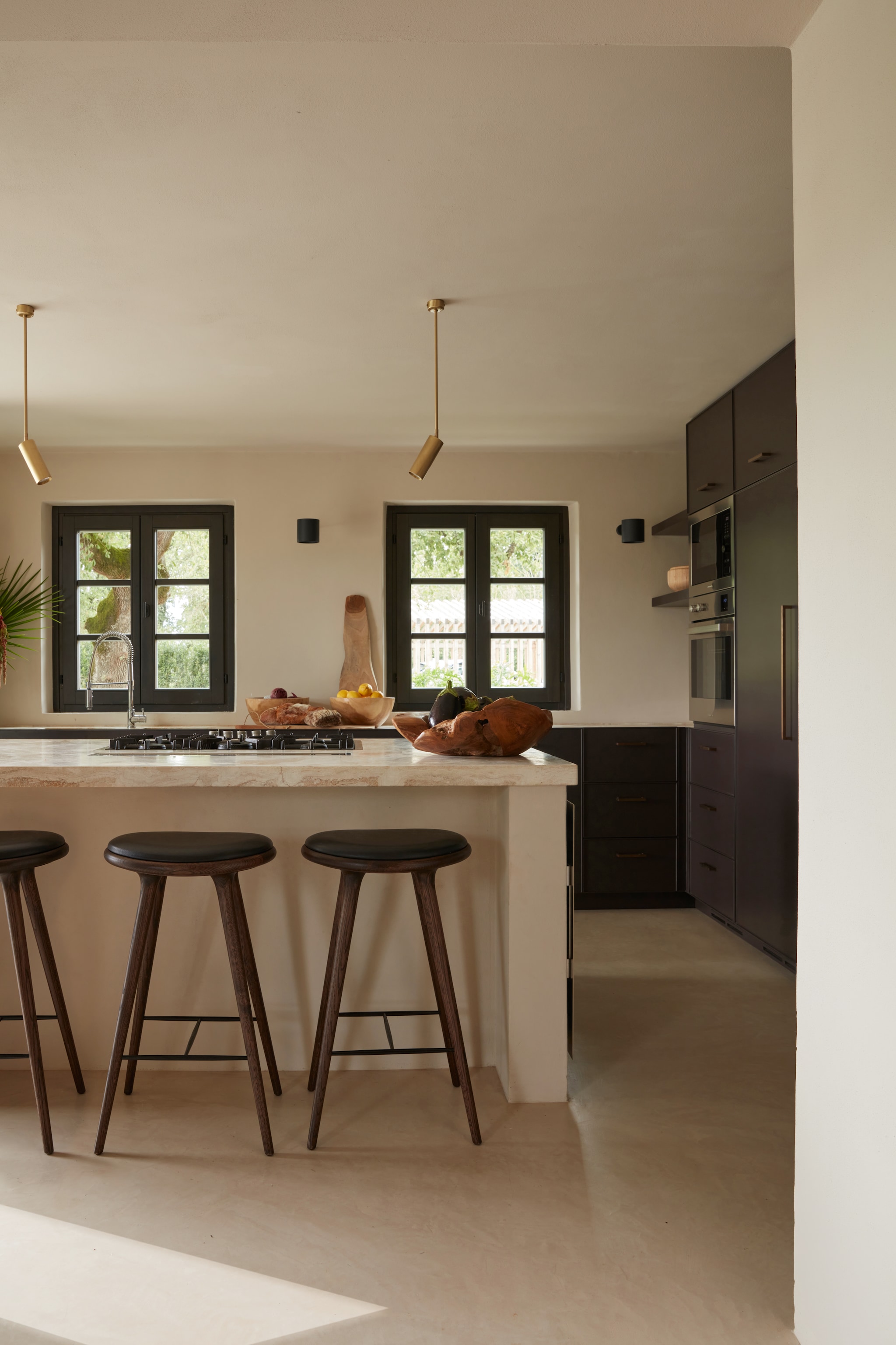 De keuken is maatwerk, keukeneiland afgewerkt met een blad van wit travertijn van Sitem Rapolano. Barkrukken High Stool van Mater. Vergelijkbare lampen via Nordic Nest. vtwonen Landelijk 07-2025 | fotografie Gaelle Le Boulicaut