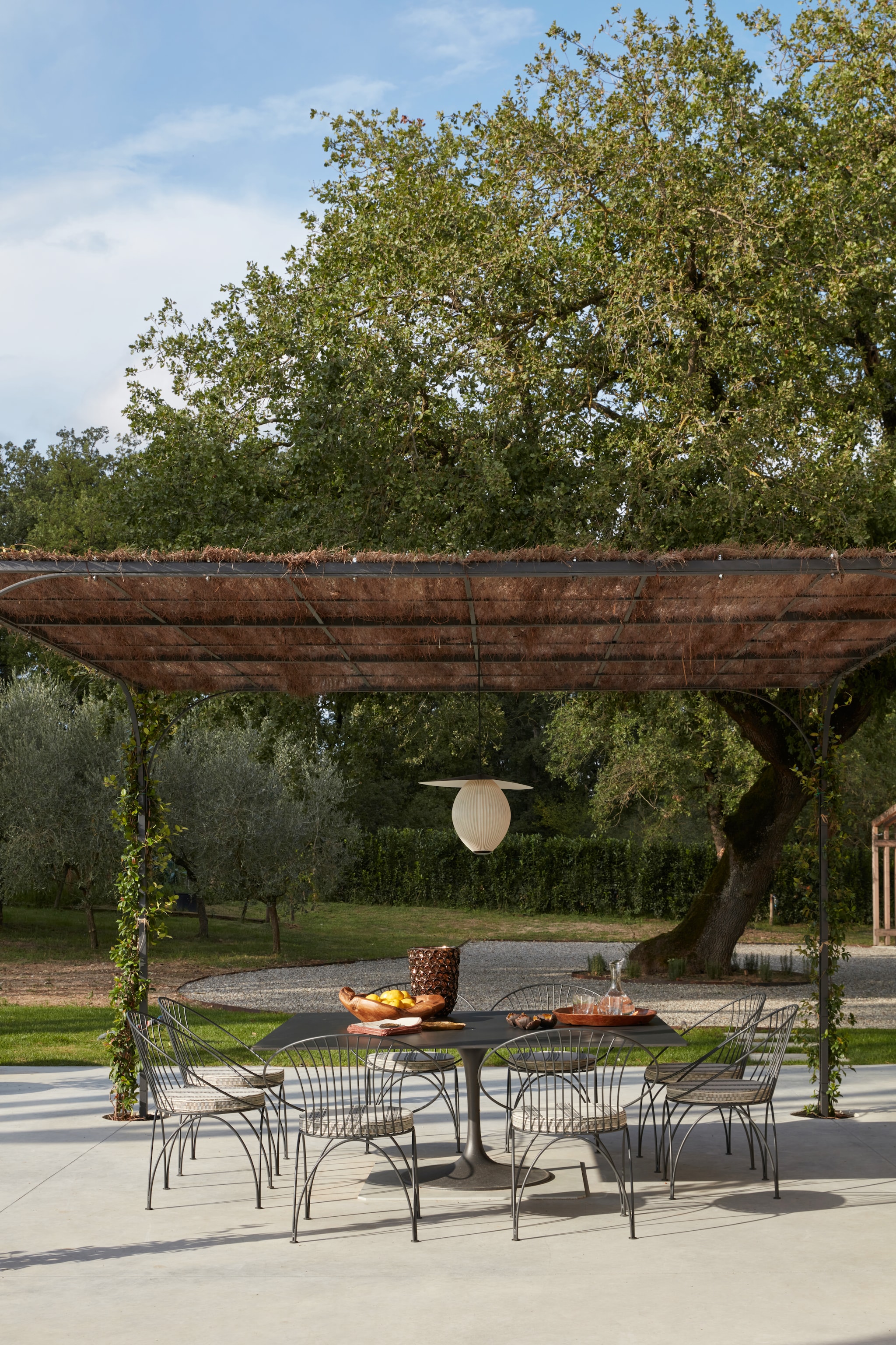 Onder de ijzeren pergola van LF Italy staat een op maat gemaakte eettafel met stoelen, ontworpen door de architect, Anne Herzog. De kussens van Paolo Nesi zorgen voor een stoere, zachte balans, met als speels accent de Satellite-lampen van Gubi. vtwonen Landelijk 07-2025 | fotografie Gaelle Le Boulicaut