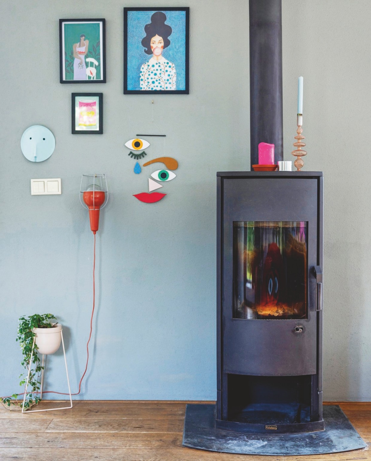 Wandkleur Pigeon van Farrow &amp; Ball. Kachel onbekend. Oranje looplamp Hema. Kunstmasker Paper Character van Lex Pott. Plantenpot Radijs Conceptstore. Mobile gezicht Voorlinden shop. Klein schilderij Etsy, twee grotere schilderijen Ali Express. Kaarsenstandaard Anna + Nina. vtwonen 05-2025 | styling Ilona Jongepier | fotografie Dana van Leeuwen