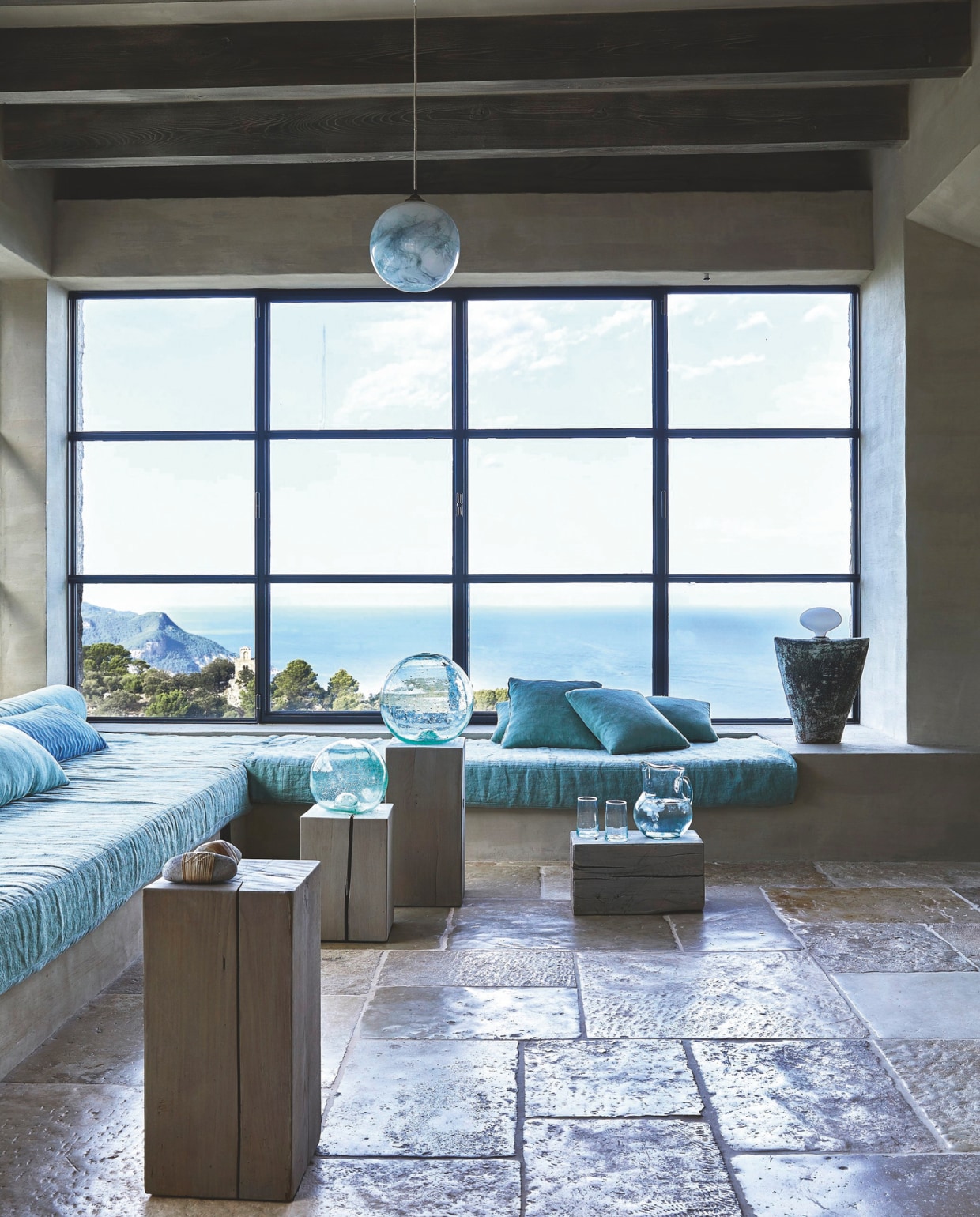 Op de vloer ligt Bourgondische steen, afkomstig uit een steengroeve in Frankrijk. De ingebouwde banken zijn bewust laag. De banken zijn bekleed met antiek Mallorcaans linnen, door More Decor met de hand geverfd met natuurlijke pigmenten om de tinten van zeeschuim en olijfbladeren te benaderen. De donkerdere blauwe kussens zijn gemaakt van antieke Japanse kusaki-zome stoffen. Gemarmerde glazen plafondlampen van een glasblazer uit Engeland en houten bijzettafels op maat gemaakt. Lamp links ontworpen door More Decor. Vazen, kan en drinkglazen van gerecycled glas van Viveca in Palma. Stijlvol Wonen 04-2025 | fotografie Greg Cox | tekstbewerking Anneleen Peeters | productie Sven Alberding/Bureaux/Living Inside | brontekst Robyn ALexander