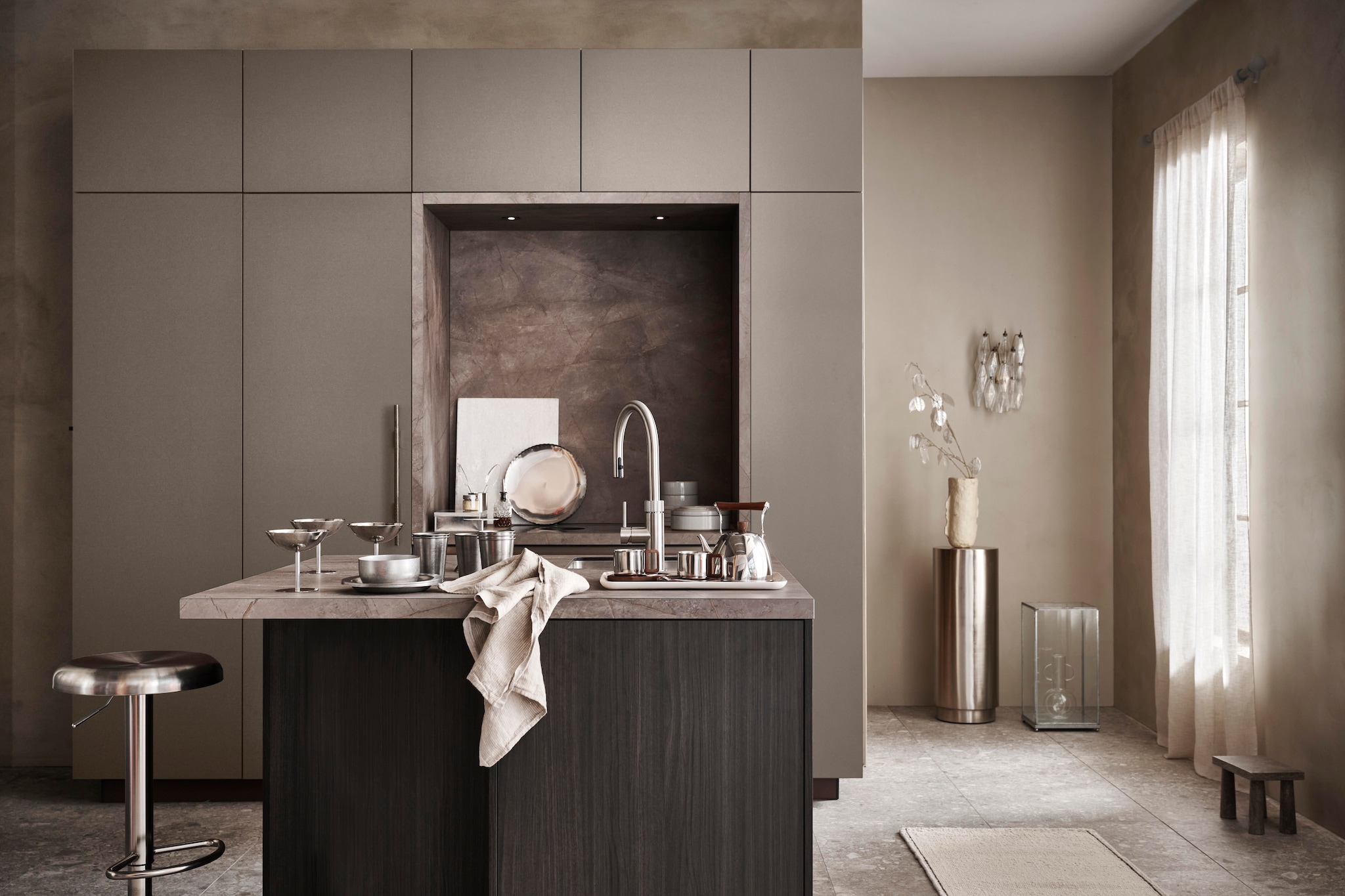 vtwonen keuken Tricolore, kwartsietgrijs, in deze opstelling € 23.500,-. M.m.v. shop.vtwonen.nl (vloertegel Composite donkergrijs), basiclabel.nl (barkruk Isaac), hetkabinet.nl (marmeren snijplank, rond schilderbord, tak met zilveren blaadjes, metalen vitrinebox, glazen vaas, beige vaas op pilaar), zarahome.com (gordijnen, travertin kruk, vloerkleed, stalen mok met acaciahouten schotel, stalen theepot met acaciahouten details, metalen wijnglazen, glazen slabestek), instagram.com/theartoflumi (glazen wandlamp), thefinestore.com (By Mölle theedoek Pinstripe Flax, Redecker metalen opbergbox, Ferm Living rvs kom, bord en bekers Tumbled, Hasami grijs porselein servies, kommen en borden, Fog Linen Work dienblad Linen Tray Natural). Privébezit (pilaar). vtwonen 07-2025 | styling Kim van Rossenberg | fotografie Sjoerd Eickmans