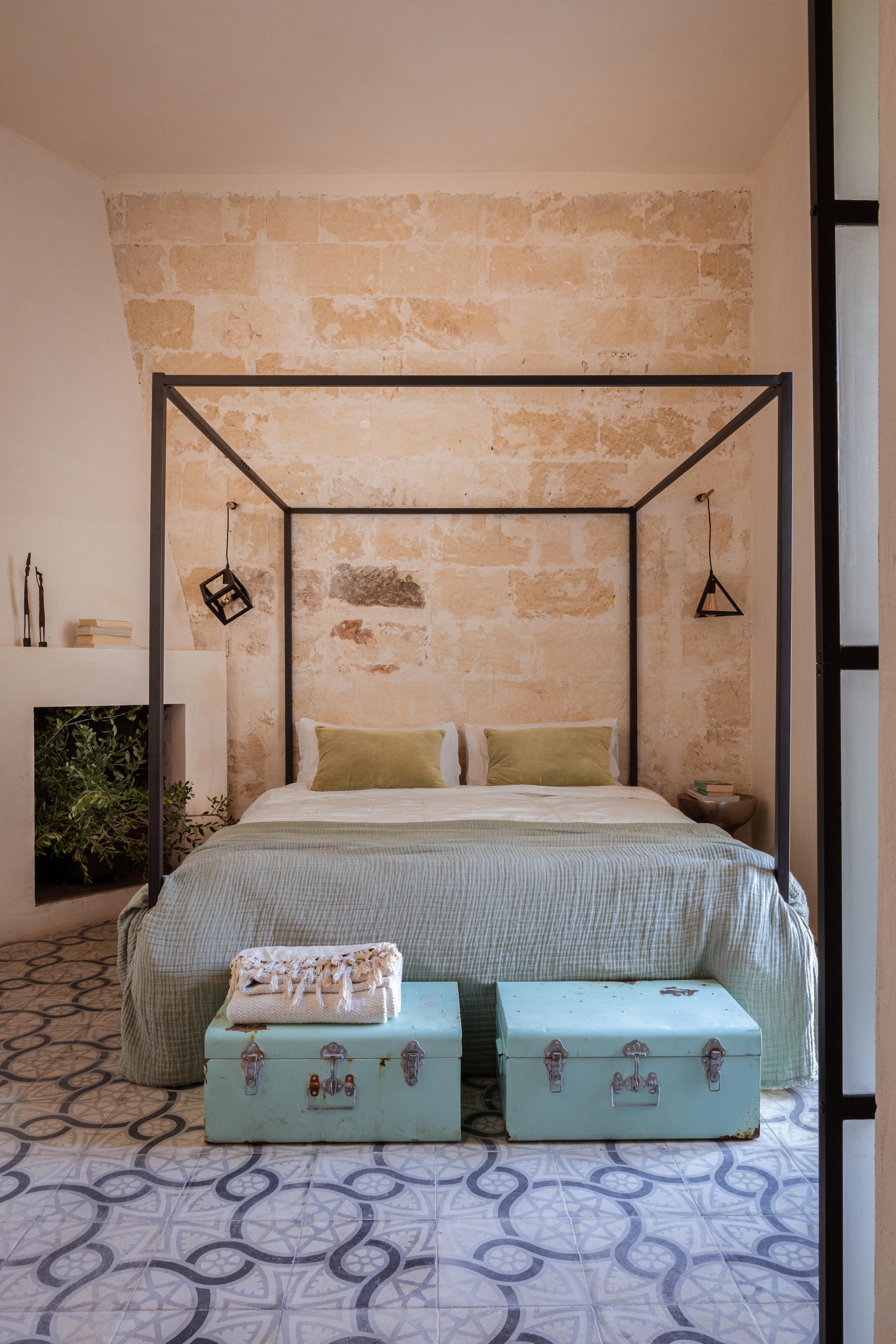 Mangohouten ladekast uit Indonesië. Hemelbed en smeedijzeren wandlampen Olenka Interiors. Fuwelen kussens H&M Home. Linnen dekbedovertrek linnen AM.PM van La Redoute. Linnenmix sprei uit Portugal. Metalen koffers vintage. Vloer antieke cementtegels. Plaid Maisons du Monde. vtwonen 08-2025 | productie Living Inside | fotografie Monica Spezia