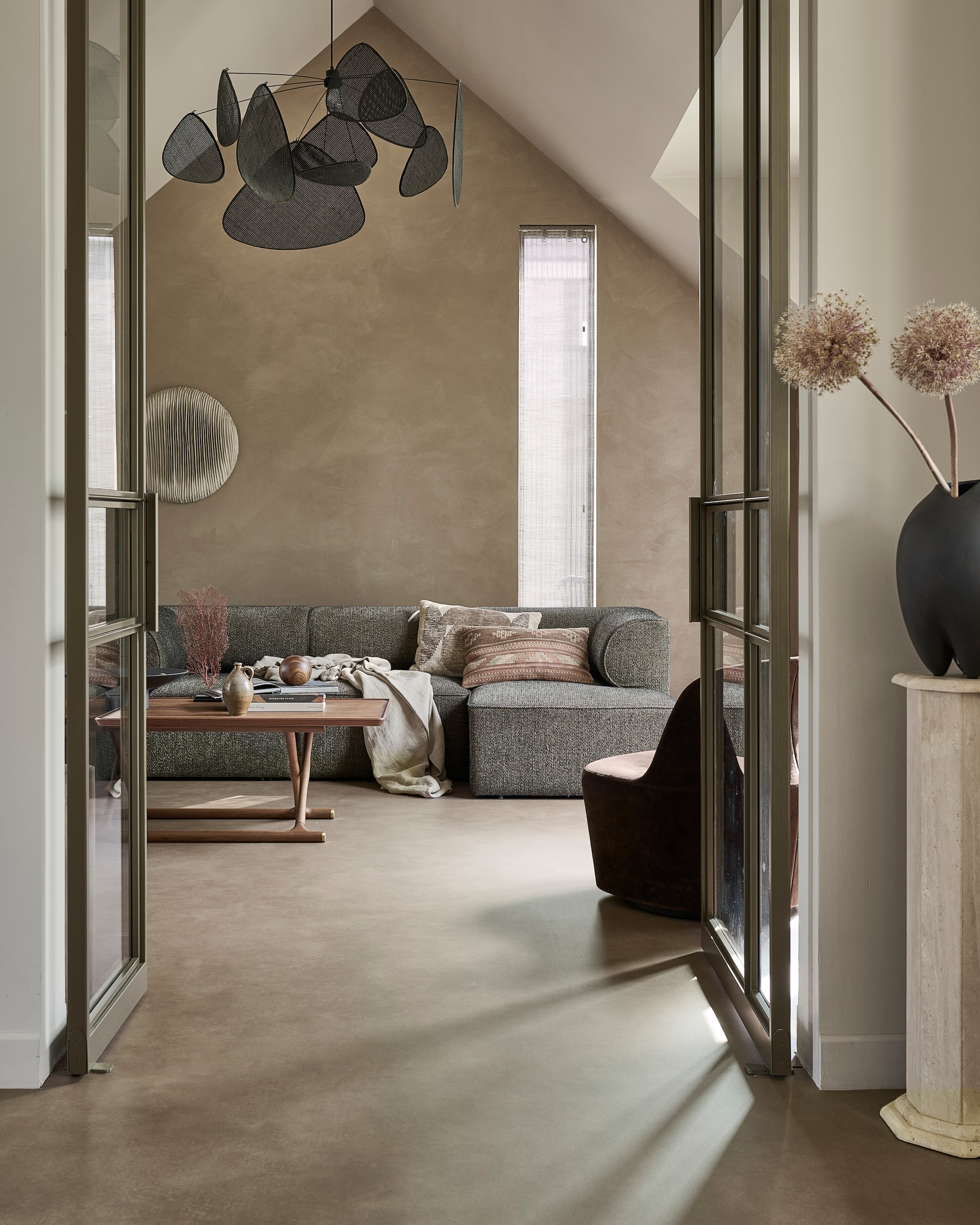 vtwonen Novilon Pumice, bronze (2118), per m² € 36,75 (exclusief leggen).M.m.v. designtales.nl (modulebank, fauteuil, salontafel), zarahome.com (plaid), 101cph.com (zwarte vaas), marketset.fr (hanglamp), skygate.nl (deuren), zonnelux.nl (raambekleding), studioastriddodd.com (wandobject). Overig privébezit. vtwonen 08-2025 | styling Kim van Rossenberg | fotografie Sjoerd Eickmans