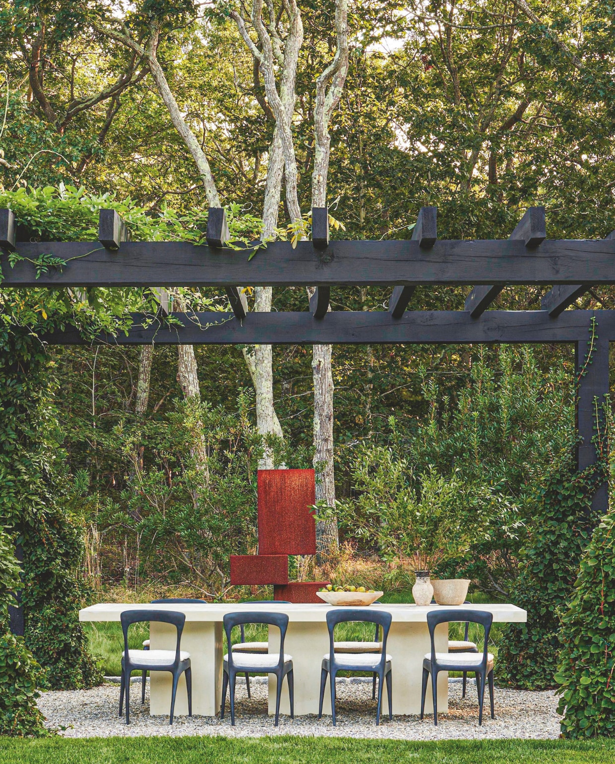 Van de ruim 7000 m² tuin was een groot deel bos. Nu zijn er verschillende zitplekken en is er ruimte voor kunst. Een terras van grind onder de zwartgebeitste pergola vormt een fijne eetplek. Stoelen Flow van Henry Hall. Betonnen tafel Carlyle Collective. Kunst Stacked van Luis Urribarri.&gt; vtwonen Landelijk 05-2025 | styling Colin King | fotografie Adrian Gaut  | tekstbewerking Margreet van Schaik