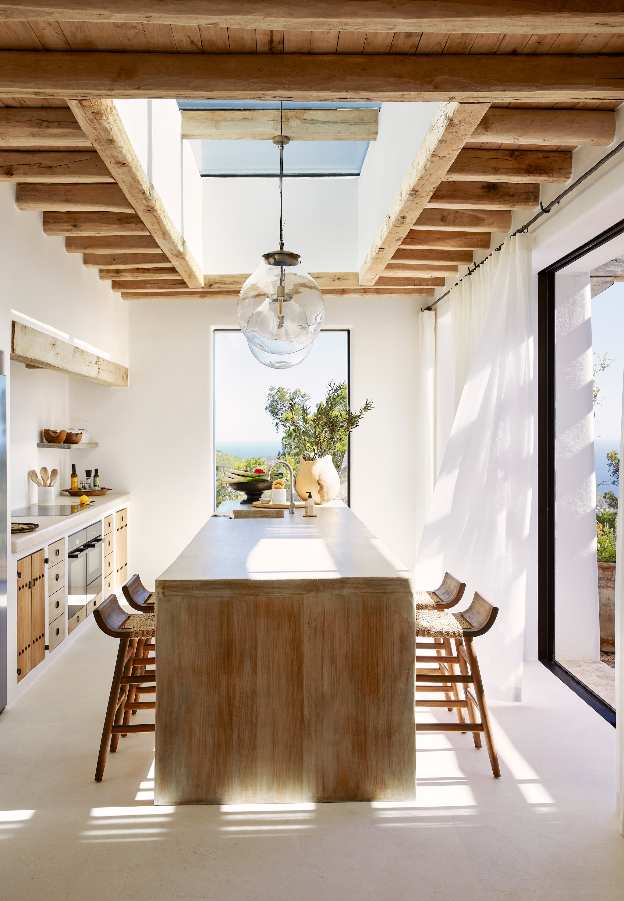 De maatwerk keuken combineert beton met houten kasten in de stijl van Ibiza. Houten vaas en schaal La Grange Interiors. Glazen lampen uit India. Op de vloer grote tegels van kalksteen van het eiland. vtwonen Landelijk 08-2025 | fotografie Elsa Young | productie Living Inside