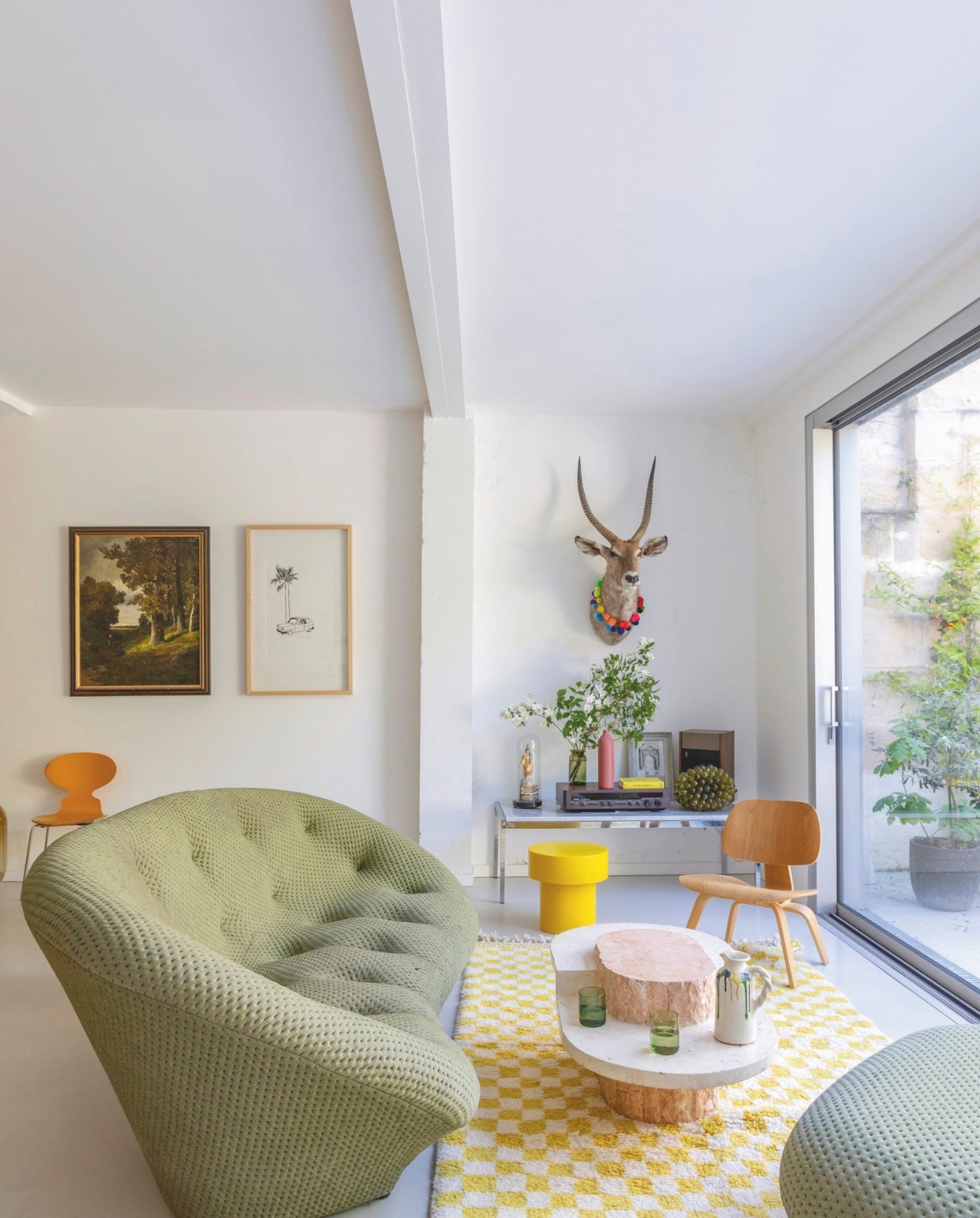 Bank Ploum van Ligne Roset, vloerkleed Berber Chico Obi Home, salontafel Maitland-Smith, stoel rechts Eames Lcw van Vitra, kruk Pion van Ohm Studio, tafel tegen wand van Florence Knoll via Pamono, hertenkop Thierry Féret. vtwonen 06-2025 | productie AC Scoffoni | styling Marie-Maud Levron | fotografie Cécile Perrinet Lhermitte