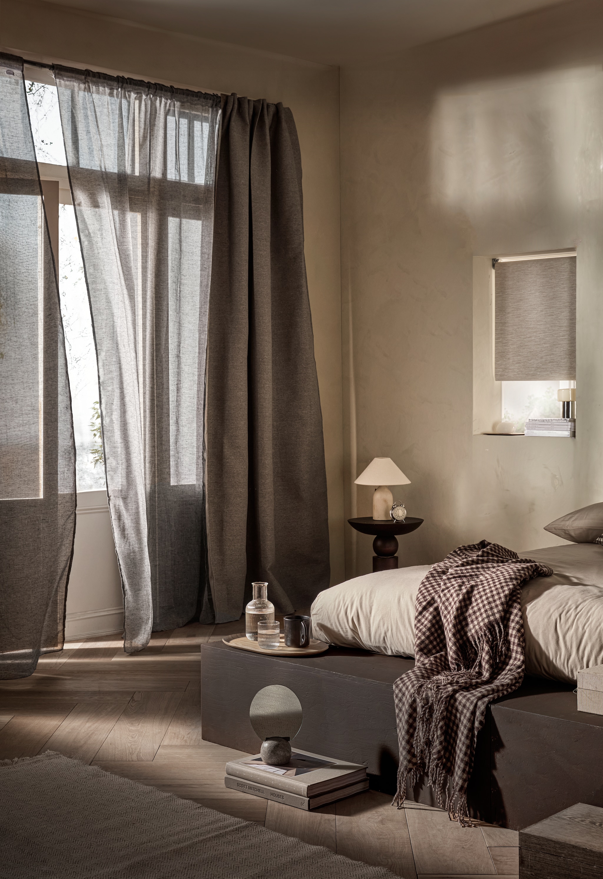 Verduisterend rolgordijn Sand, 60x190 cm (bxh) € 83,99, verduisterend gordijn Drizzle in Mirre, met plooi,  140x270 cm (bxh) € 73,99 en halftransparant gordijn Frost in Grijs, met plooi, 140x270cm (bxh) € 50,99.M.m.v. vestingh.nl (kalkverf Chalk van Lab in Just Jimmy), zarahome.com (tafellamp, bijzettafel, wekker, karaf met glas, plaid, kleine tafellamp kozijn, ronde albasten tafellamp, dienblad), hm.com (mok, spiegel op voet, dekbedovertrek). Overig privébezit. vtwonen 06-2025 | styling Sanne Ketelaar | fotografie Peggy Janssen