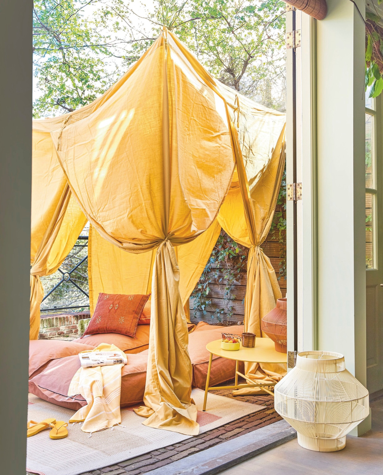 Katoenen klamboe € 550,- (hkliving.com). Waterafstotend daybed Strech € 650,- (bryck.nl). Kussen in oranje € 44,95, katoenen hammamdoek geel € 13,95 (rozenkelim.nl). Kunststof buitenkleed Paxi € 29,99, metalen buitentafel Chris in geel € 39,99 (karwei.nl). Gerecycled plastic lampionlamp No2 S in Terra € 114,95, gerecycled plastic lampionlamp No2 M in Naturel € 169,95 (originalhome.nl). Keramieken kopjes  en schaal (evafollendergrossfeld.nl). Slippers van Havaianas € 22,- (thelittlegreenbag.nl). Overig privébezit. vtwonen 06-2025 Styling Hello Yellow | styling Kelly van Gendt | fotografie Peggy Janssen