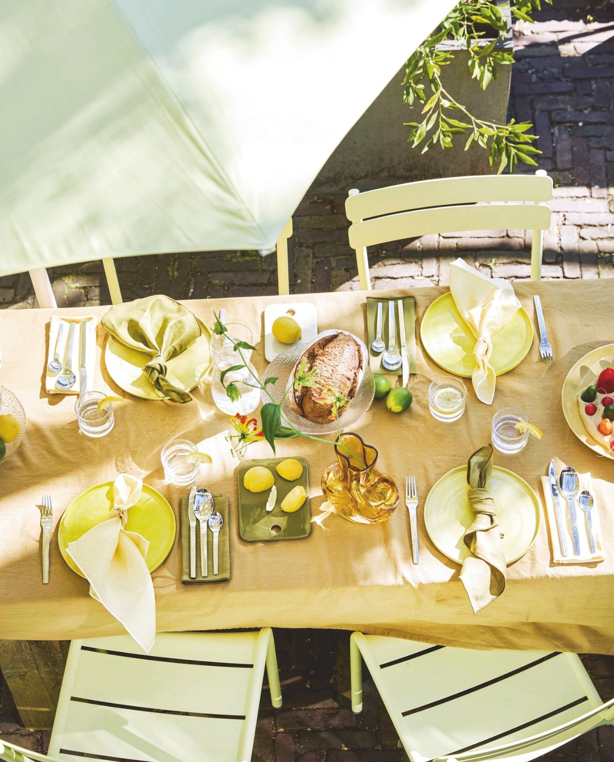 Aluminium tuinstoel Luxembourg van Fermob in Frosted Lemon € 269,-, parasol van Fermob in Frosted Lemon € 455,-, aluminium vogelvoederhuisje Picoti van Fermob in Frosted lemon € 65,- (extravert.nl). Katoenen tafelkleed Washed Mosterd van Kook € 39,99 (bol.com). Glazen vaas Gleam € 24,95 (hkliving.com). Linnen servetten van Kulu Club in olijfgroen en botergeel, set van 2 € 22,95, glazen waterkan Sui Bubbles Clear van Hübsch € 22,-, glazen Sui van Hübsch € 6,-, katoenen theedoek in bruin van Hübsch € 12,-, keramieken borden Amare in olijfgroen van Hübsch € 38,-, stalen bestek Mano van Hübsch € 100,-, glazen kom Fuyu van Hübsch € 8,50 (shop.vtwonen.nl). vtwonen 06-2025 Styling Hello Yellow | styling Kelly van Gendt | fotografie Peggy Janssen