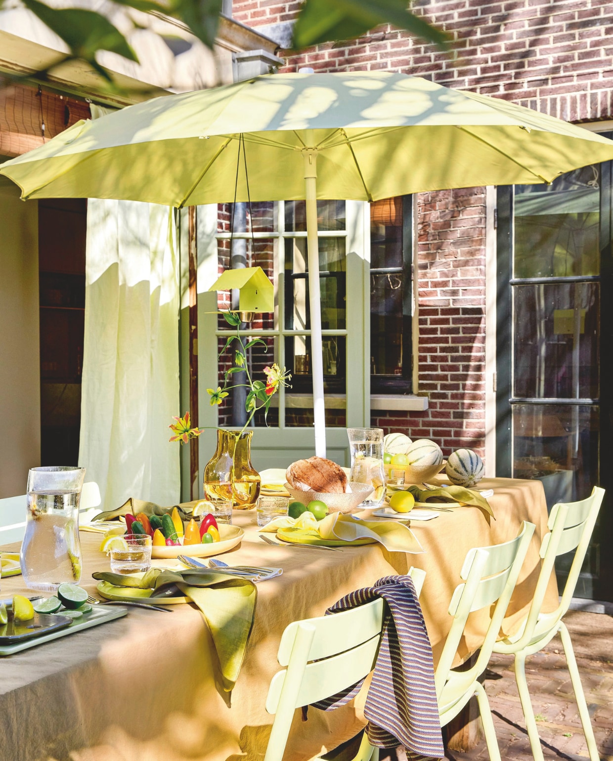 Aluminium tuinstoel Luxembourg van Fermob in Frosted Lemon € 269,-, parasol van Fermob in Frosted Lemon € 455,-, aluminium vogelvoederhuisje Picoti van Fermob in Frosted lemon € 65,- (extravert.nl). Katoenen tafelkleed Washed Mosterd van Kook € 39,99 (bol.com). Glazen vaas Gleam € 24,95 (hkliving.com). Linnen servetten van Kulu Club in olijfgroen en botergeel, set van 2 € 22,95, glazen waterkan Sui Bubbles Clear van Hübsch € 22,-, glazen Sui van Hübsch € 6,-, katoenen theedoek in bruin van Hübsch € 12,-, keramieken borden Amare in olijfgroen van Hübsch € 38,-, stalen bestek Mano van Hübsch € 100,-, glazen kom Fuyu van Hübsch € 8,50 (shop.vtwonen.nl). vtwonen 06-2025 Styling Hello Yellow | styling Kelly van Gendt | fotografie Peggy Janssen