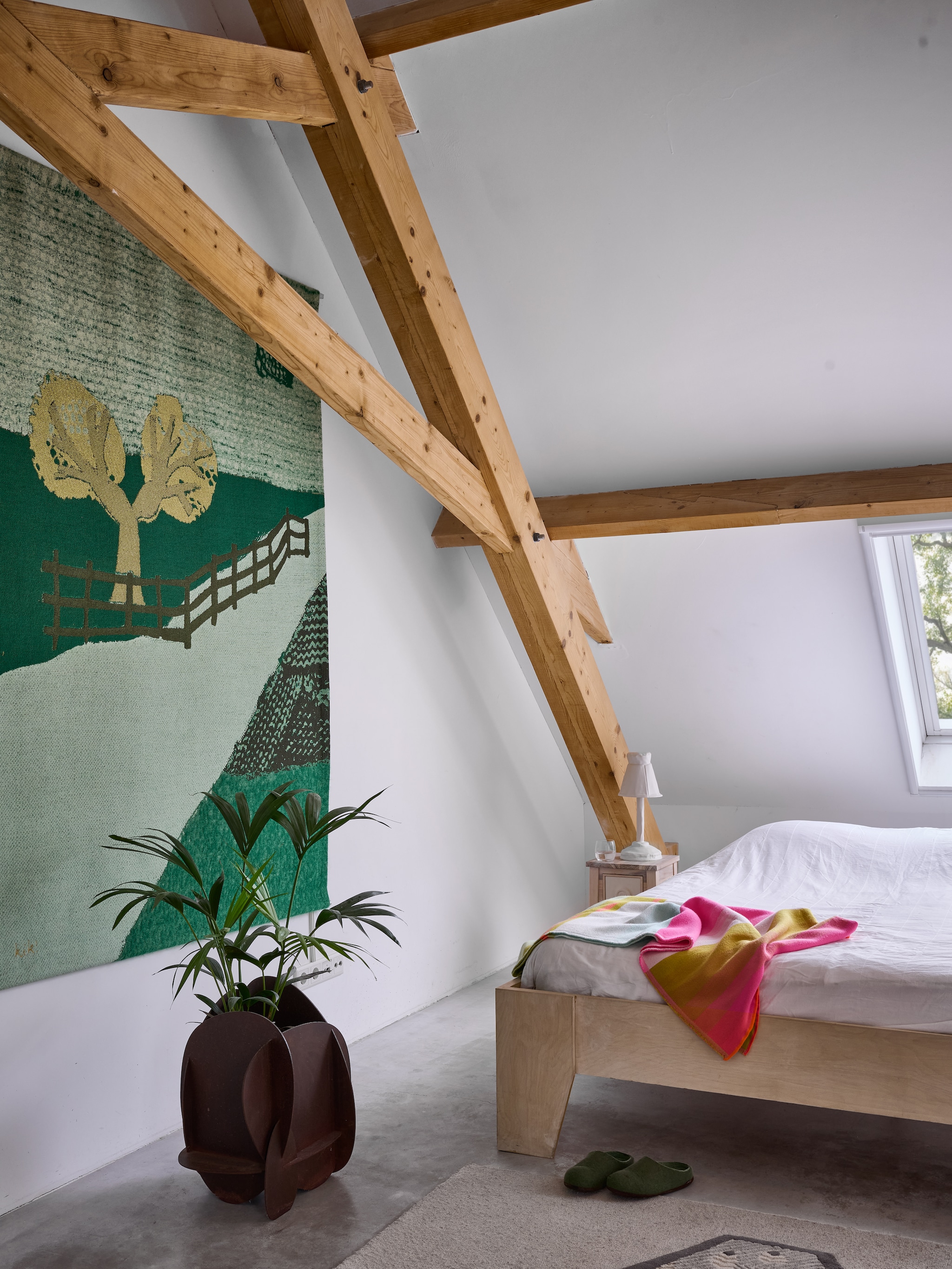 Aan de wand in de slaapkamer op de eerste verdieping een wandkleed uit de serie Memories of a Panorama van Kiki. Ervoor een indoor variant van de Interlocking Planters van cortenstaal. Op het nachtkastje een witte keramieken Soft Table Shade van Kiki. Op bed een Colour Plaid van Scholten & Baijings voor Hay. Stijlvol Wonen 8-2025 | fotografie Kasia Gatkowska