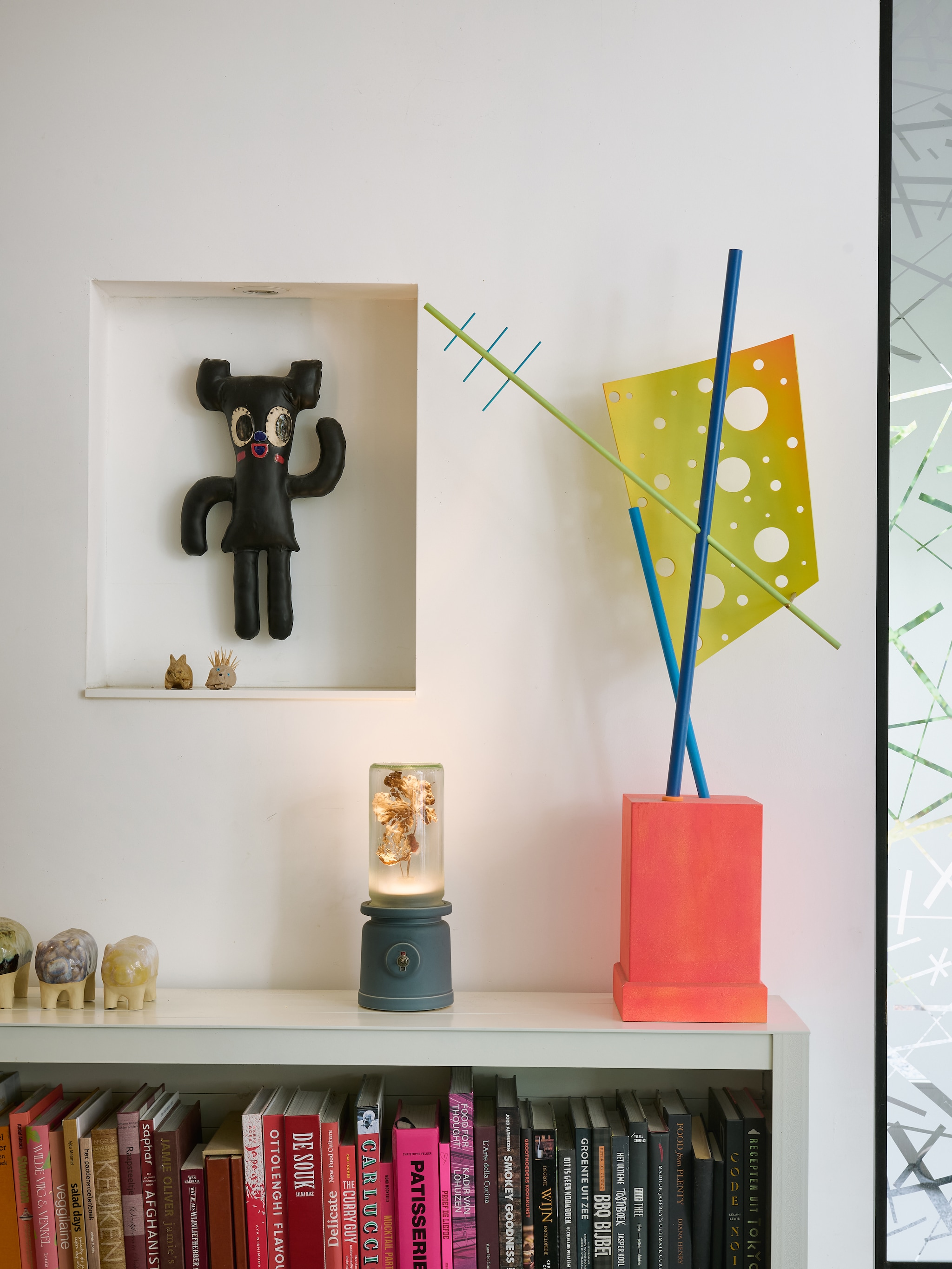 Op de boekenkast staan drie spaarvarkentjes, die oud-student Martha Nash maakte voor haar afstudeerproject aan de Design Academy Eindhoven. In het midden lamp Meck van Kranen/Gille, geproduceerd door keramiekatelier Cor Unum. Rechts daarvan een sculptuur uit de Tinker-serie van Joost. In de nis een keramieken pop van Bas Kosters, uitgegeven in samenwerking met Cor Unum. Stijlvol Wonen 8-2025 | fotografie Kasia Gatkowska