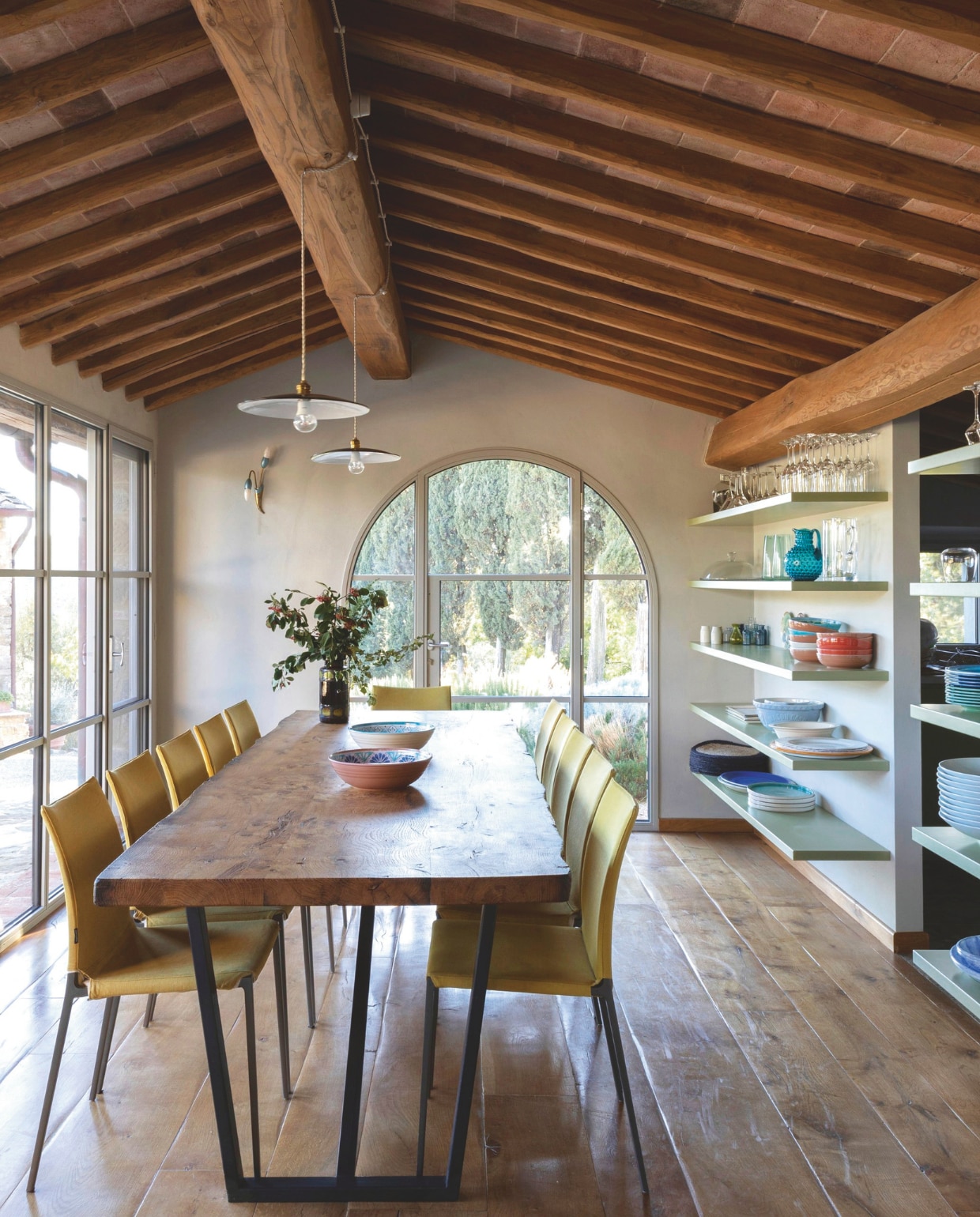 Het plafond werd gerestaureerd met kastanjehouten balken met ertussen terracotta tegels. Tafel op maatgemaakt van eiken en ijzer. Stoelen Lia van Roberto Barbieri voor Zanotta. Typisch Toscaanse hanglampen. Maatwerk schappen in mat saliegroen. vtwonen landelijk 7-2025 | productie Living Inside | fotografie Monica Spezia