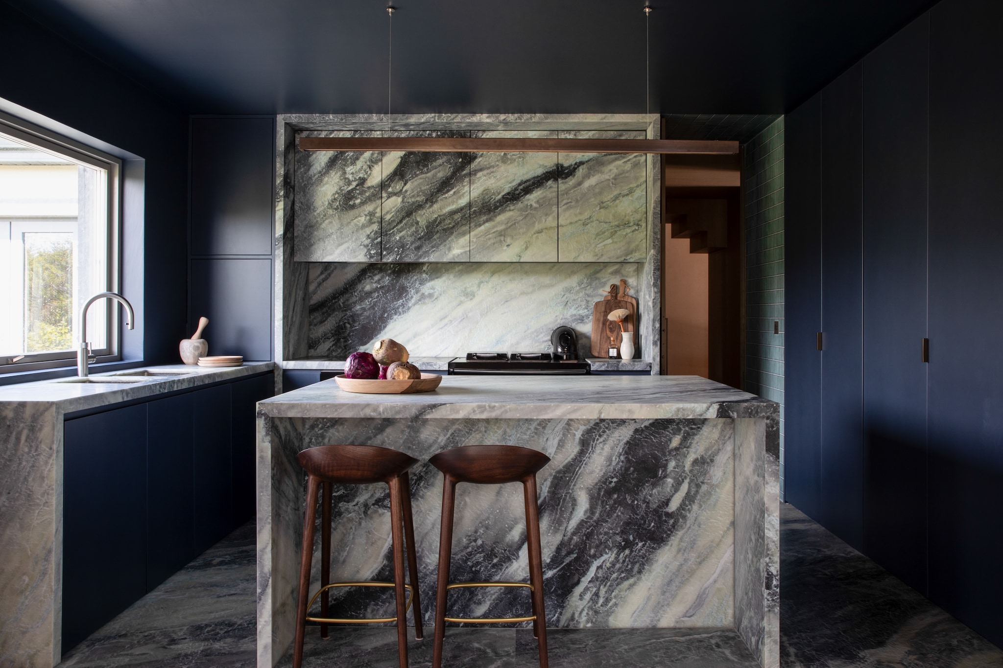 De keukenbladen en achterwand zijn vervaardigd van Seafoam-kwartsiet, een materiaal dat onder hoge druk en temperatuur is samengeperst en zeer kras- en hittebestendig is. Barkrukken Inoda + Sveje. Hanglamp Anour, uitgevoerd in bruin koper. Stijlvol Wonen 10-2025 | fotografie Ruth Maria Murphy