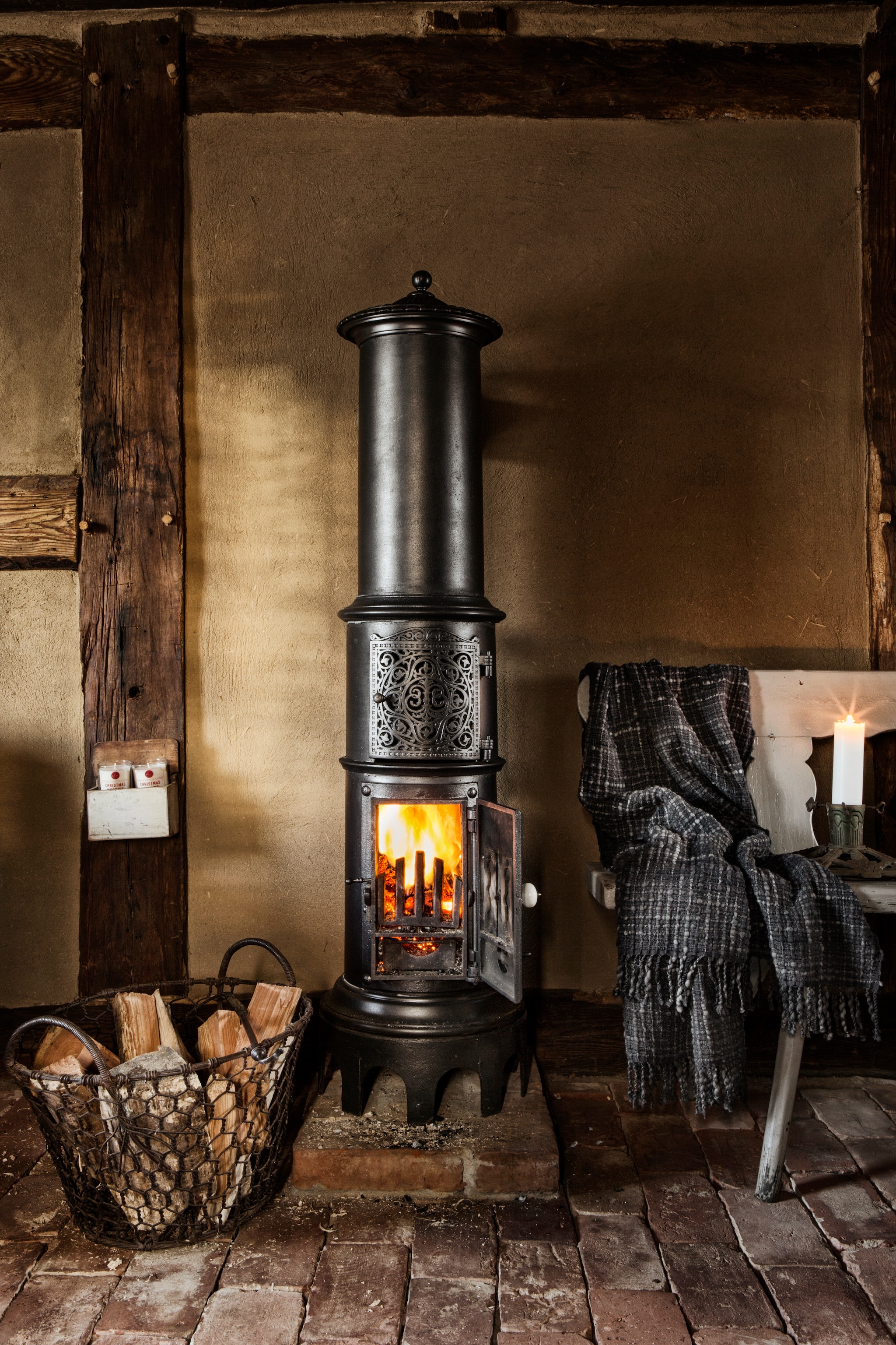 Haard 19de-eeuwse Deense kolomkachel. De handgemaakte wollen plaid was een cadeau. Antieke kerstboom-standaard met een  witte altaarkaars en manden Unikat Depot. vtwonen landelijk 11-2025 | productie The Country House | styling Miriam Hannemann | fotografie André Reuter