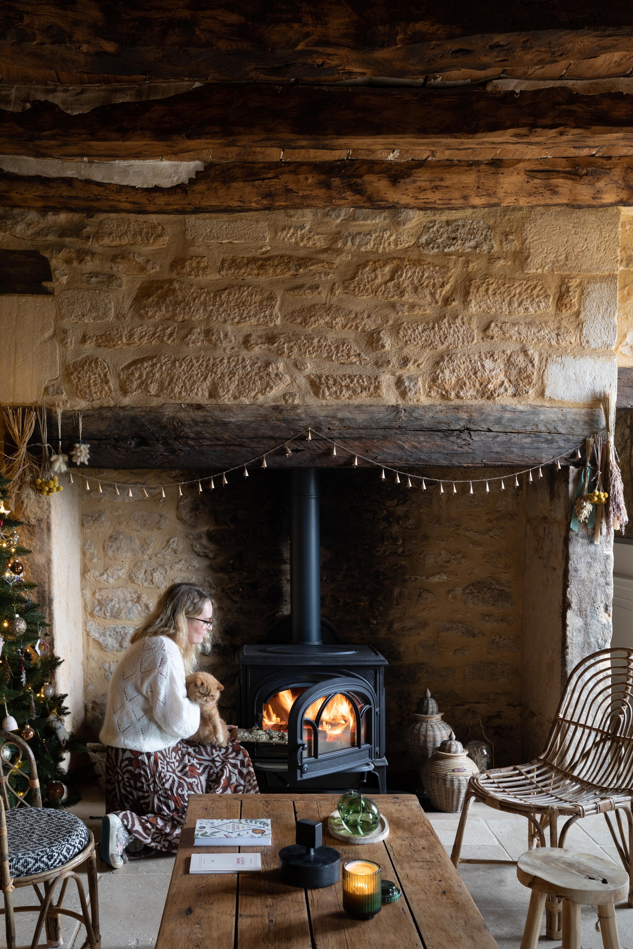 De massieve open haard (cheminée cantou) is typerend voor de architectuur van het huis. De bewoners plaatsten er een houtkachel in, wat voor meer praktische warmte zorgt. Potten Rivièra Maison. Slinger uit India. Salontafel en kruk rommelmarkt. Bamboe stoel Madam Stoltz. vtwonen Landelijk 11-2025 | styling Wilma Custers | fotografie Fabienne Delafraye