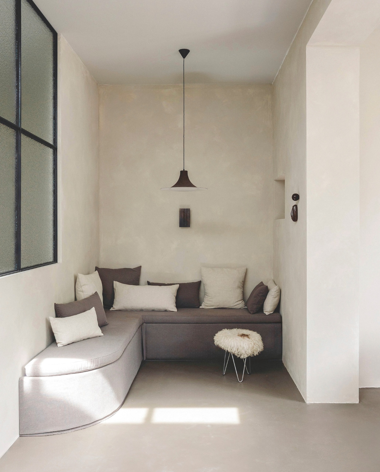 Maatwerk bank Meubels Van Zant, gestoffeerd door Noortje ter Hark. Kussens van stoffen uit Barcelona en van De Boerenbonthal. Kruk zelfgemaakt. Vintage hanglamp Gubi via Kringloop vintage & more. Wandlamp Serax. Kunstwerk Edith Beurskens & April van Domburg. Stalen raam Gillissen Ontwerp. Op de muur betonverf Silk, op de vloer beton ciré Ground Cover, beide Beton Cire Webshop. vtwonen landelijk 8-2025 | fotografie Stie De Neve