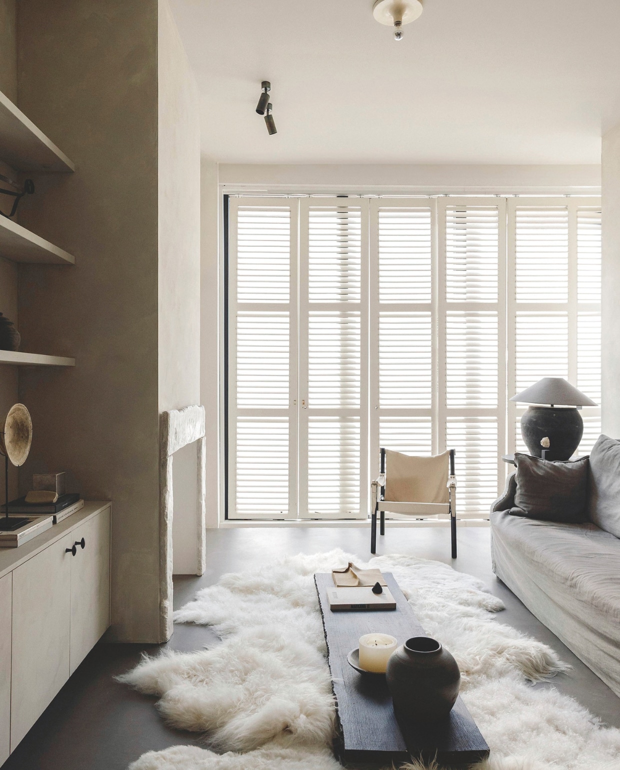 Bank en kussen Marktplaats. Stoel Kringloop Vintage & more, frame zelf zwart gespoten. Schouw Rustic Stones Schouwen. Vintage shutters Mammoet Oude Bouwmaterialen. Lamp van keramieken pot en ronde tafel Luksa Home Collection. Kleed van schapenvachten en lage, zwarte salontafel zelfgemaakt. Keramieken object kringloop Antwerpen. Bruine pot antiek. Kandelaar Marktplaats. vtwonen landelijk 8-2025 | fotografie Stie De Neve