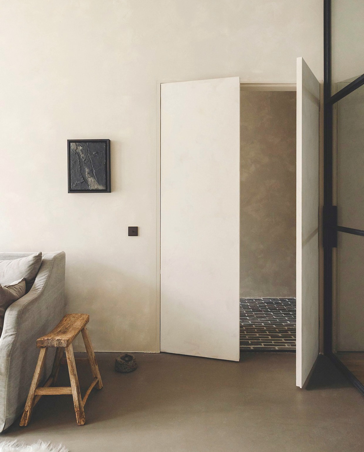 Deuren geverfd met betonverf in Silk van Beton Cire Webshop. Staal met glazen wand Gillissen Ontwerp. Kunstwerk Atelier Earth. Houten kruk Luksa Home Collection. Stenen deurstopper vintage. Lichtschakelaar schakelwip cilinder in dark gelakt aluminium van Jung. vtwonen landelijk 8-2025 | fotografie Stie De Neve