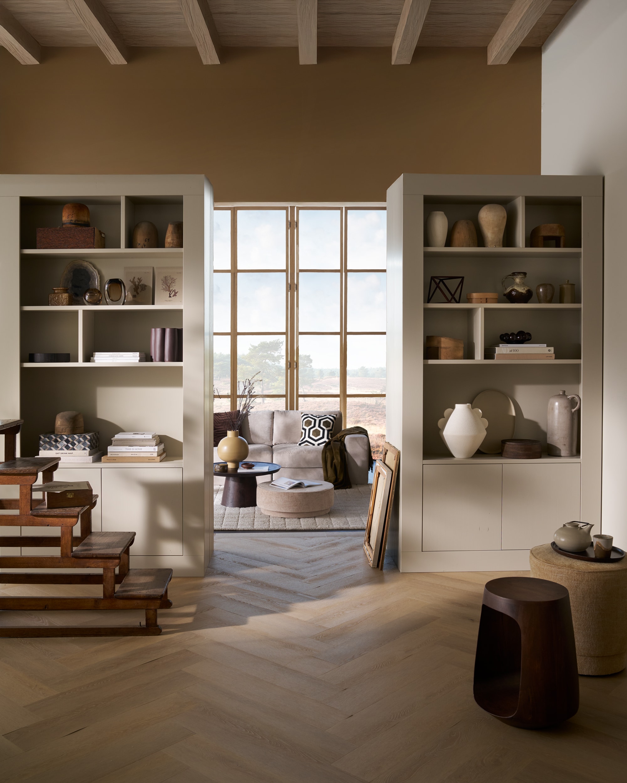 vtwonen wandkast Wallcover, grenen, dust, 117x31x229 cm (bxdxh), per stuk € 799,-, vtwonen poef Pearl en poef Pearl XL, melange stof, naturel, 40x45 cm (Øxh) € 199,- en 80x28 cm (Øxh)€ 199,-, vtwonen salontafel Coffee A Gogo, mangohout, walnoot, 90x40 cm (Øxh) € 499,- en vtwonen 2,5-zitselementenbank Family, geweven stof, zand, 298x98x85 cm (bxdxh) € 1999,-.M.m.v. pietjonker.nl (bibliotheektrap, bijzettafel), hm.com (grafisch kussen), weldaad.com (servies, opbergmand, linnen kussens), burbriantiek.nl (kruiken), vtwonen.nl (visgraat laminaat Rafter, vloerkleed Blocks). Overig privébezit.