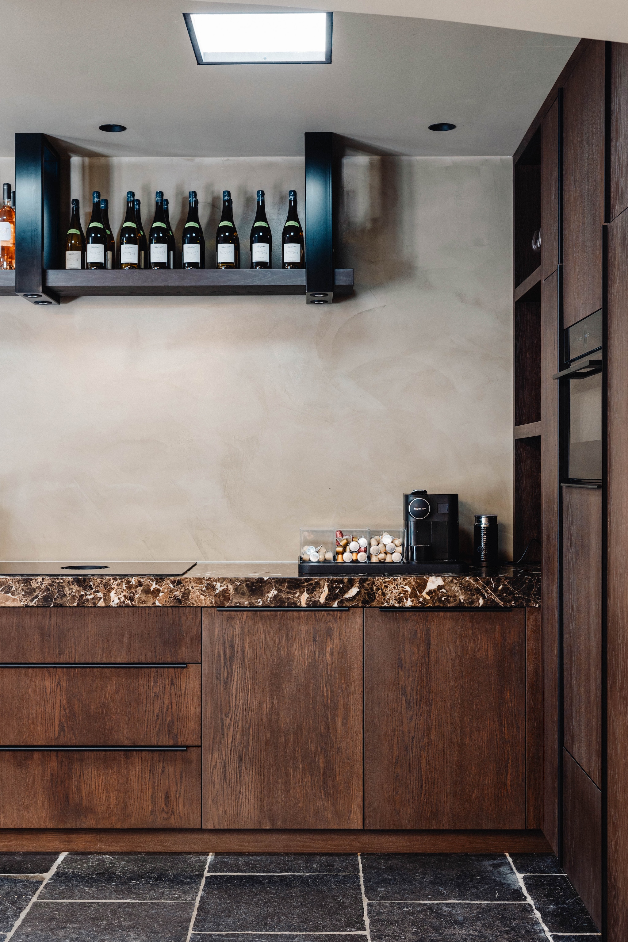 Het wijnrek in de keuken van de werkruimte is op maat gemaakt van donker hout en steekt mooi af tegen de wand van beton ciré. Stijlvol Wonen 9-2025 | fotografie Stie de Neve