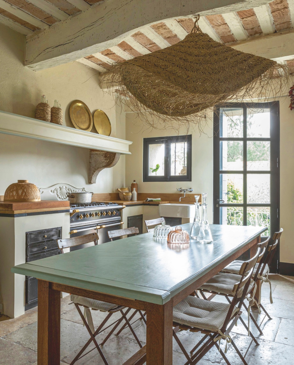 De keuken met een tafel geschikt voor twaalf personen. Het blad is geschilderd in Uzès-groen. AGA-fornuis met messing knoppen. Lampenkap van stro en een verzameling antieke kristallen flessen. vtwonen Landelijk 8-2025 | styling Laura Taccari | fotografie Monica Spezia | tekstbewerking Roel Janssen