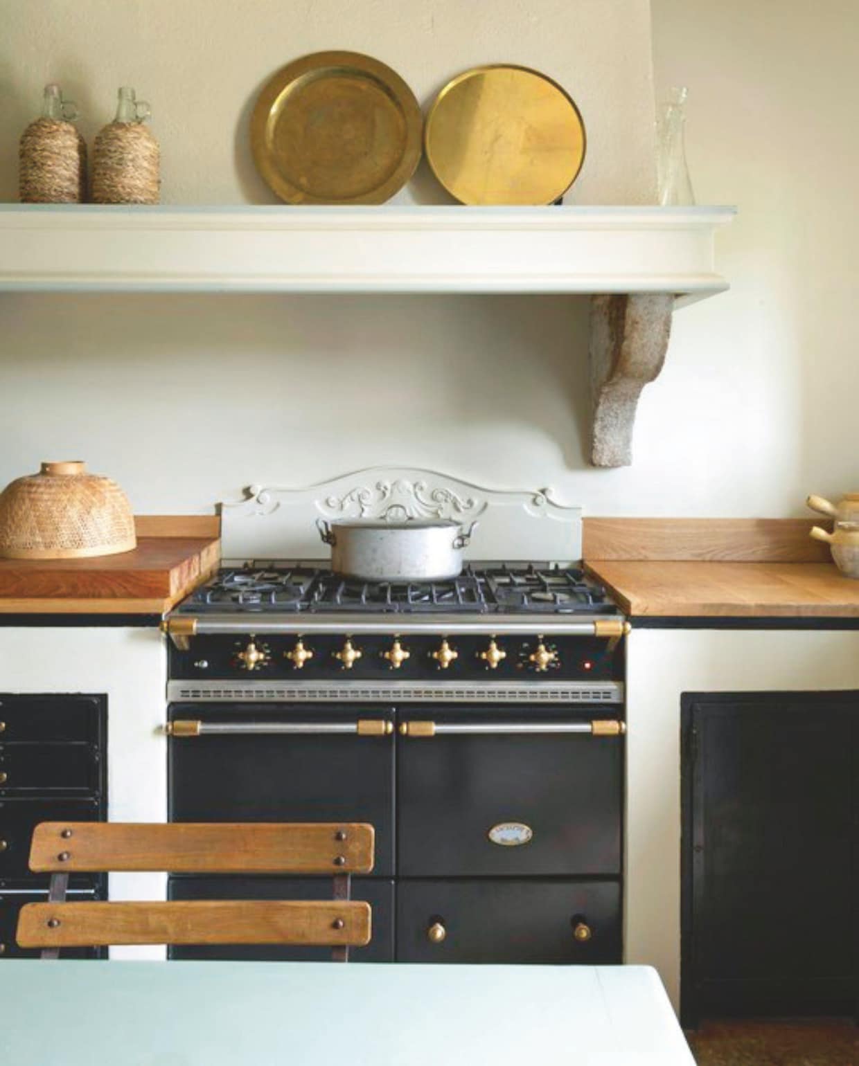 De keuken met een tafel geschikt voor twaalf personen. Het blad is geschilderd in Uzès-groen. AGA-fornuis met messing knoppen. Lampenkap van stro en een verzameling antieke kristallen flessen. vtwonen Landelijk 8-2025 | styling Laura Taccari | fotografie Monica Spezia | tekstbewerking Roel Janssen