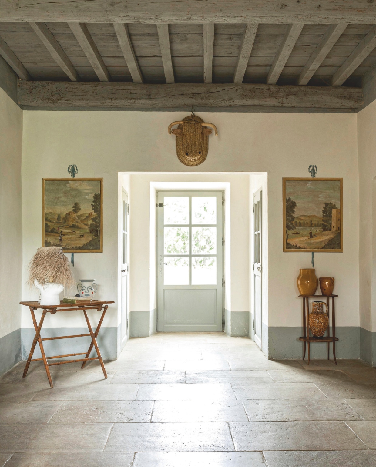 De entree van het huis. De originele vloer is van Montpellier-steen. Aan weerszijden hangen schilderijen met landelijke taferelen. De lampenkap in de vorm van een gipsen stierenkop kocht Sofie in Uzès. vtwonen Landelijk 8-2025 | styling Laura Taccari | fotografie Monica Spezia | tekstbewerking Roel Janssen